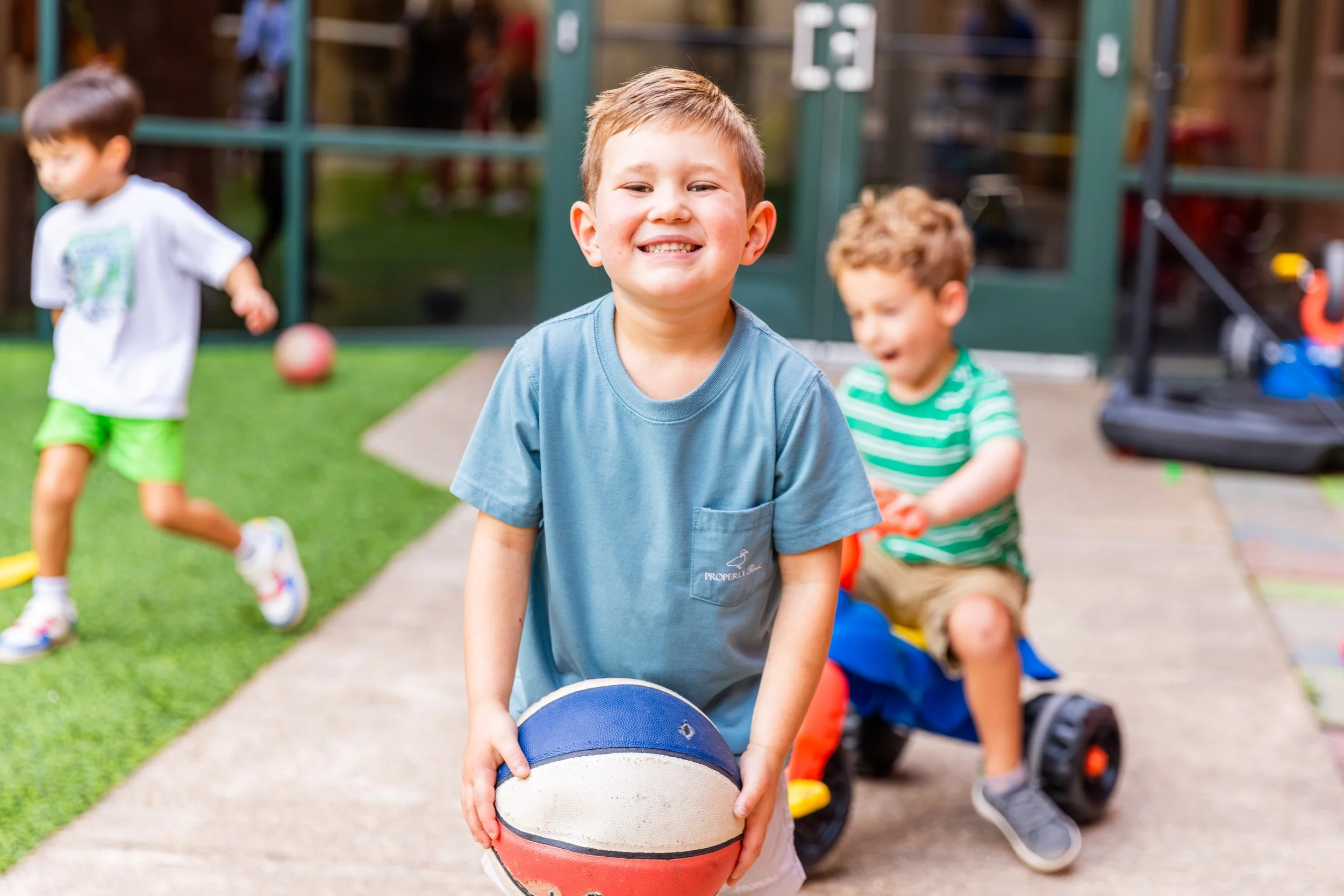 Young boy holding a basketball outdoors with children playing in the background.