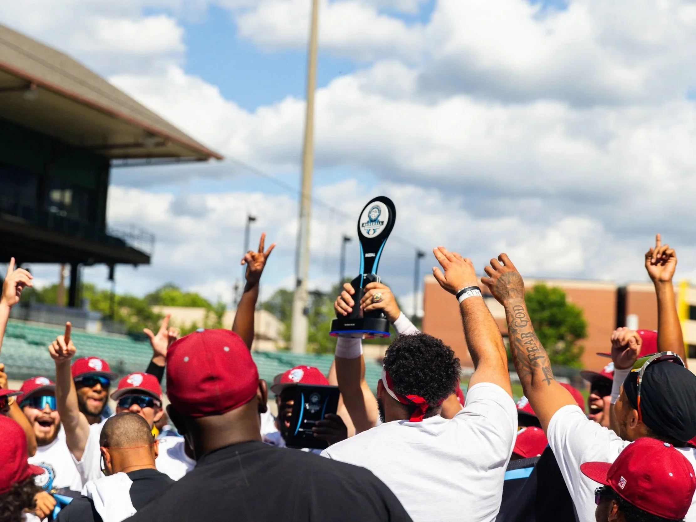 A group of sports team members celebrating outdoors, some holding a trophy, with raised hands and making victory signs, wearing red caps and sunglasses, under cloudy skies.