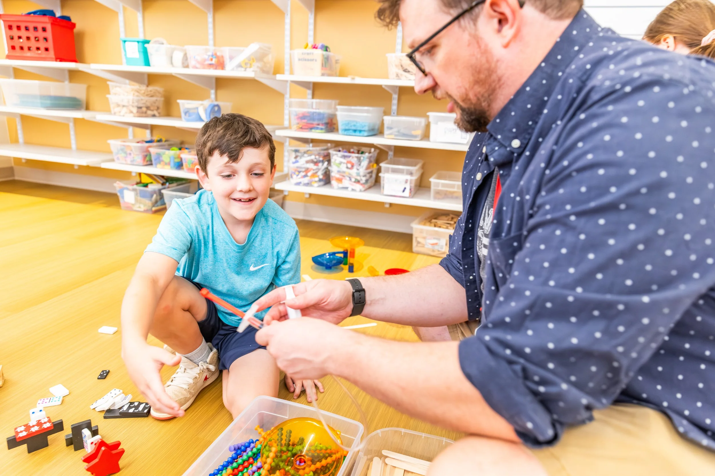 A young boy and an adult man sitting on the floor of a playroom, playing with colorful toys and building blocks.