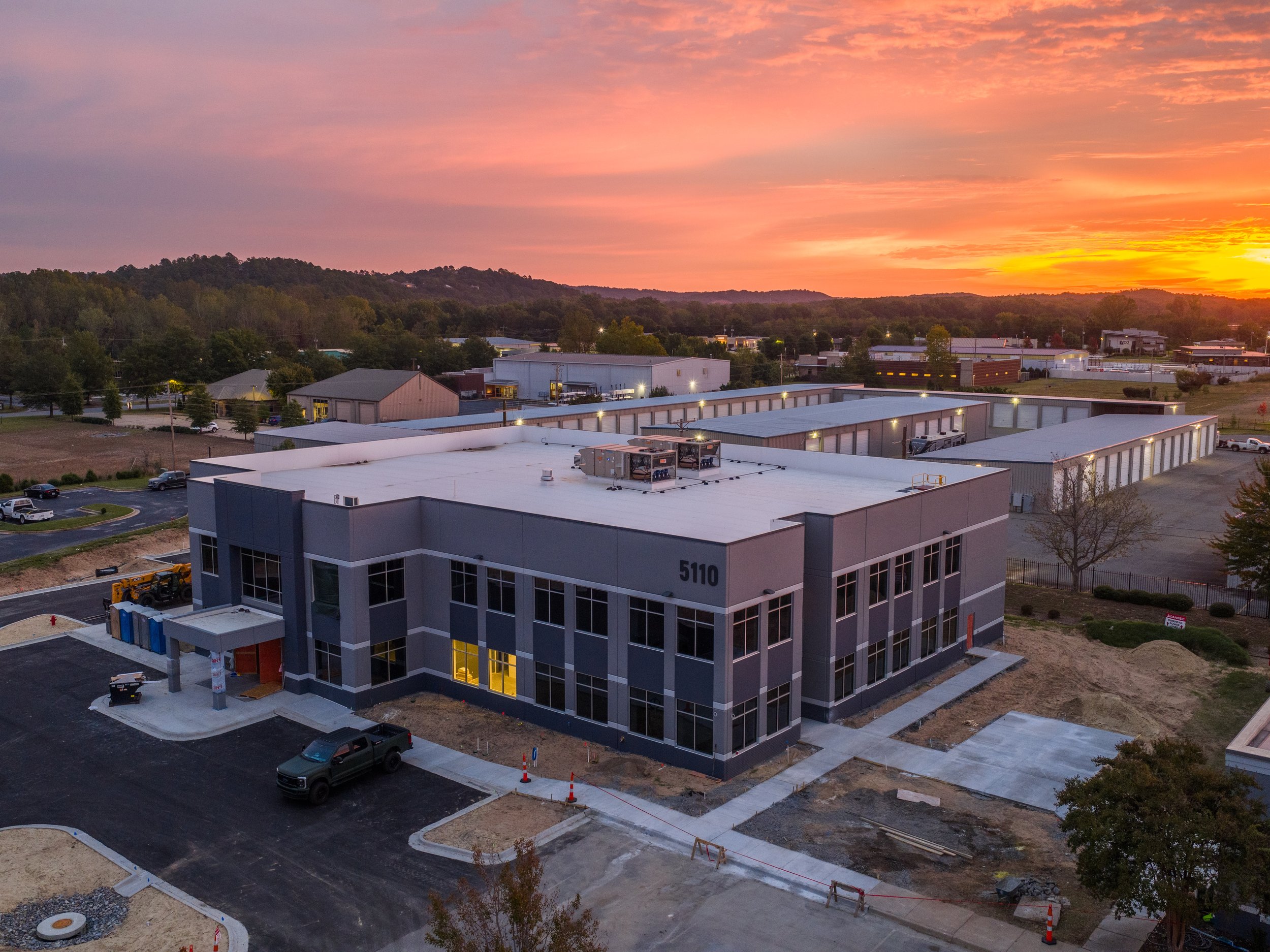 An aerial view of a commercial or industrial building complex under construction at sunset, with new parking areas, surrounding trees, and distant rolling hills.