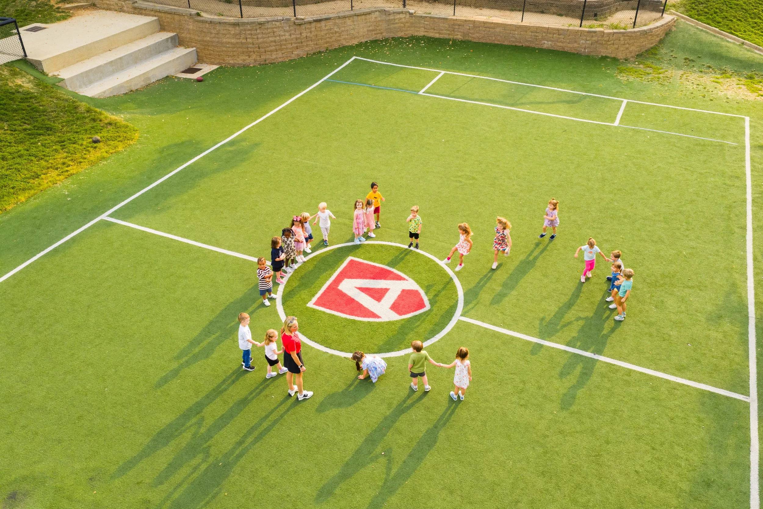 Children holding hands and forming a large circle on a green playground with white lines, around a painted logo in the center.