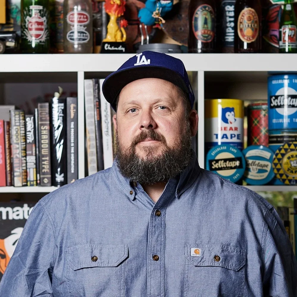Travis Price. A man with a beard wearing a blue Los Angeles Dodgers cap and a gray Carhartt shirt standing in front of a bookshelf filled with books, tapes, and decorative items.