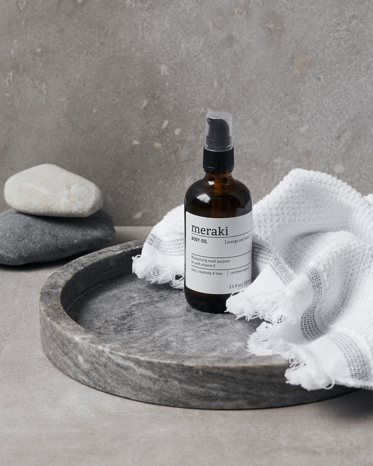 En brun glasflaska av bodyscrub med spraylock, placerad på en rund skål av sten. Runt flaskan ligger en vit, texturerad handduk, och i bakgrunden ligger två stenar, en vit och en svart, på en yta av ljusgrå material. Väggarna är samtidigt ljusgrå.
