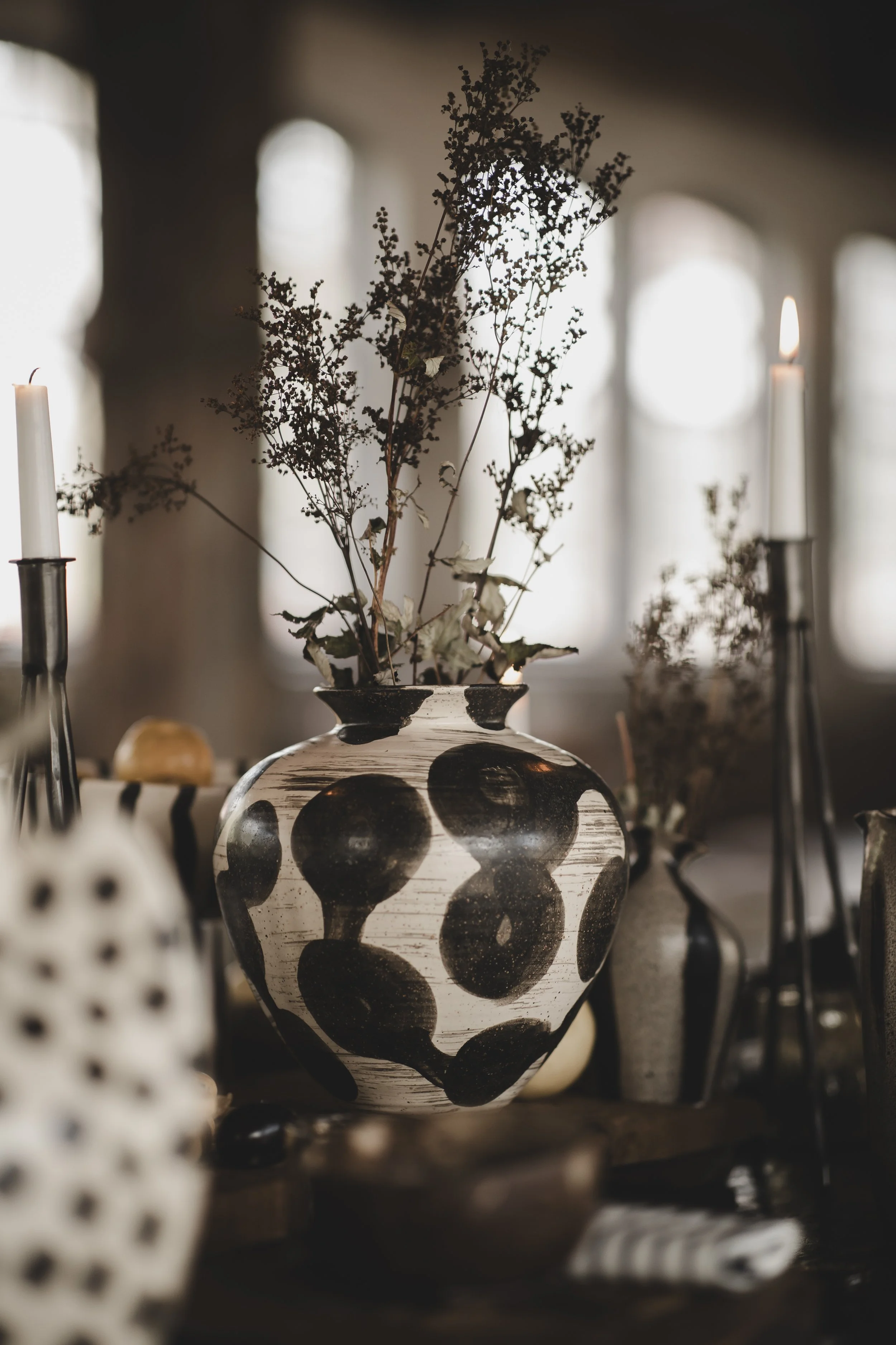 En keramikvas med svart och vit geometrisk mönster, fylld med grenar. Omkring vasan finns ljuslyktor med ljus.