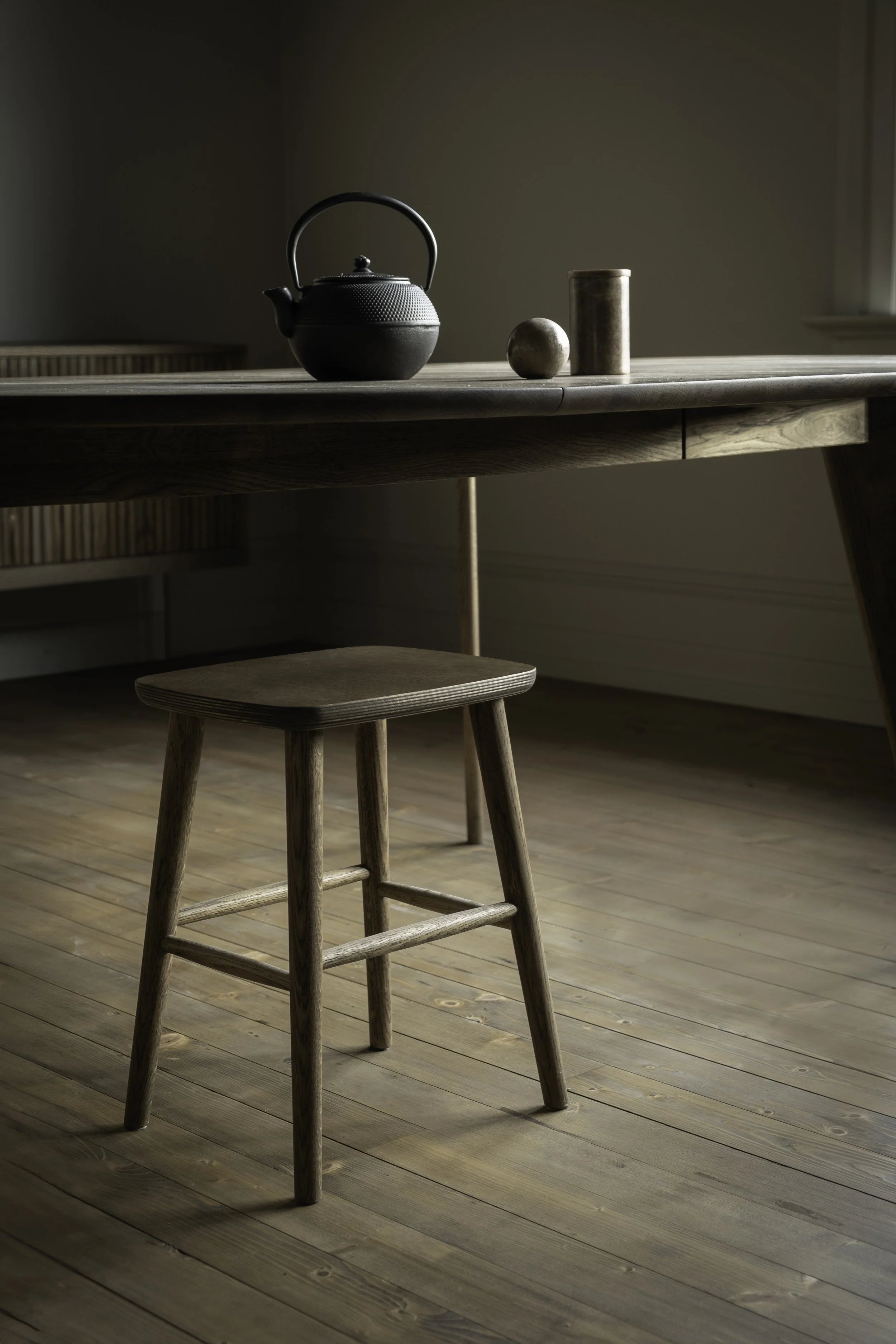 Ett minimalistiskt kök eller matsal med ett träbord och en träpall. På bordet finns en svart teekanna och tre olika små föremål, inklusive en boll och cylindrisk behållare.