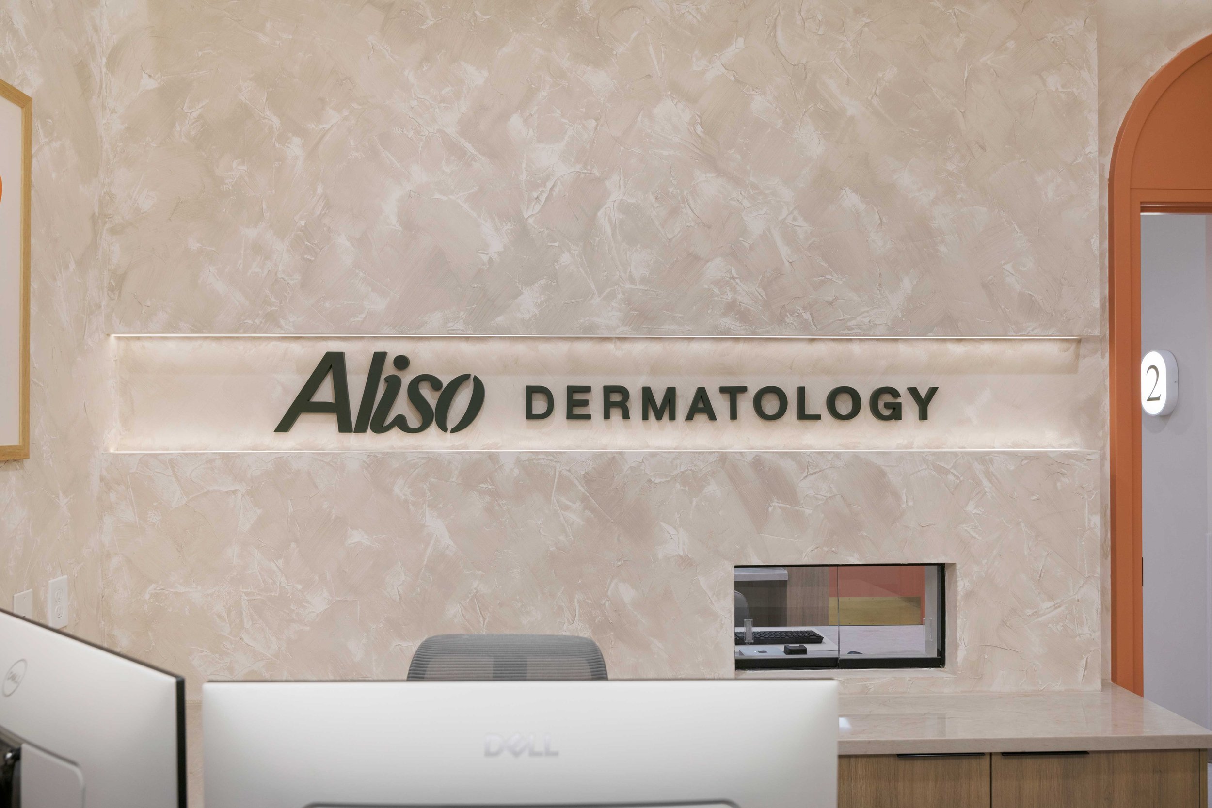 Front reception desk view at Aliso Dermatology featuring a modern recessed logo sign on a textured beige accent wall.