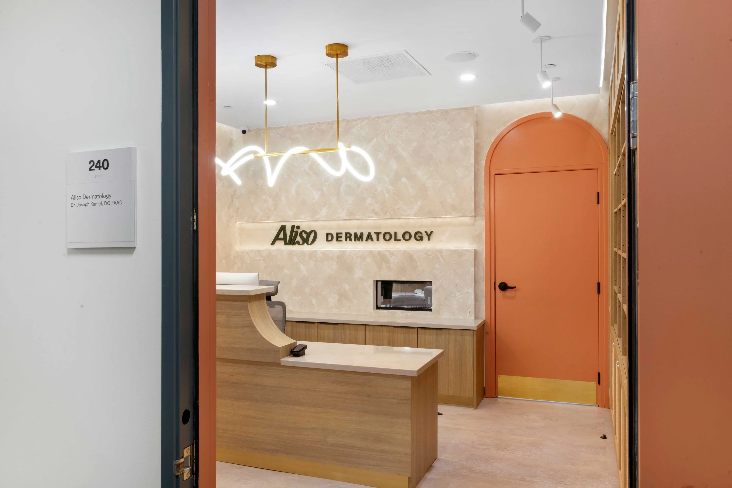 Entrance to Aliso Dermatology clinic featuring a modern reception desk, warm wood accents, and a distinctive peach arched door.