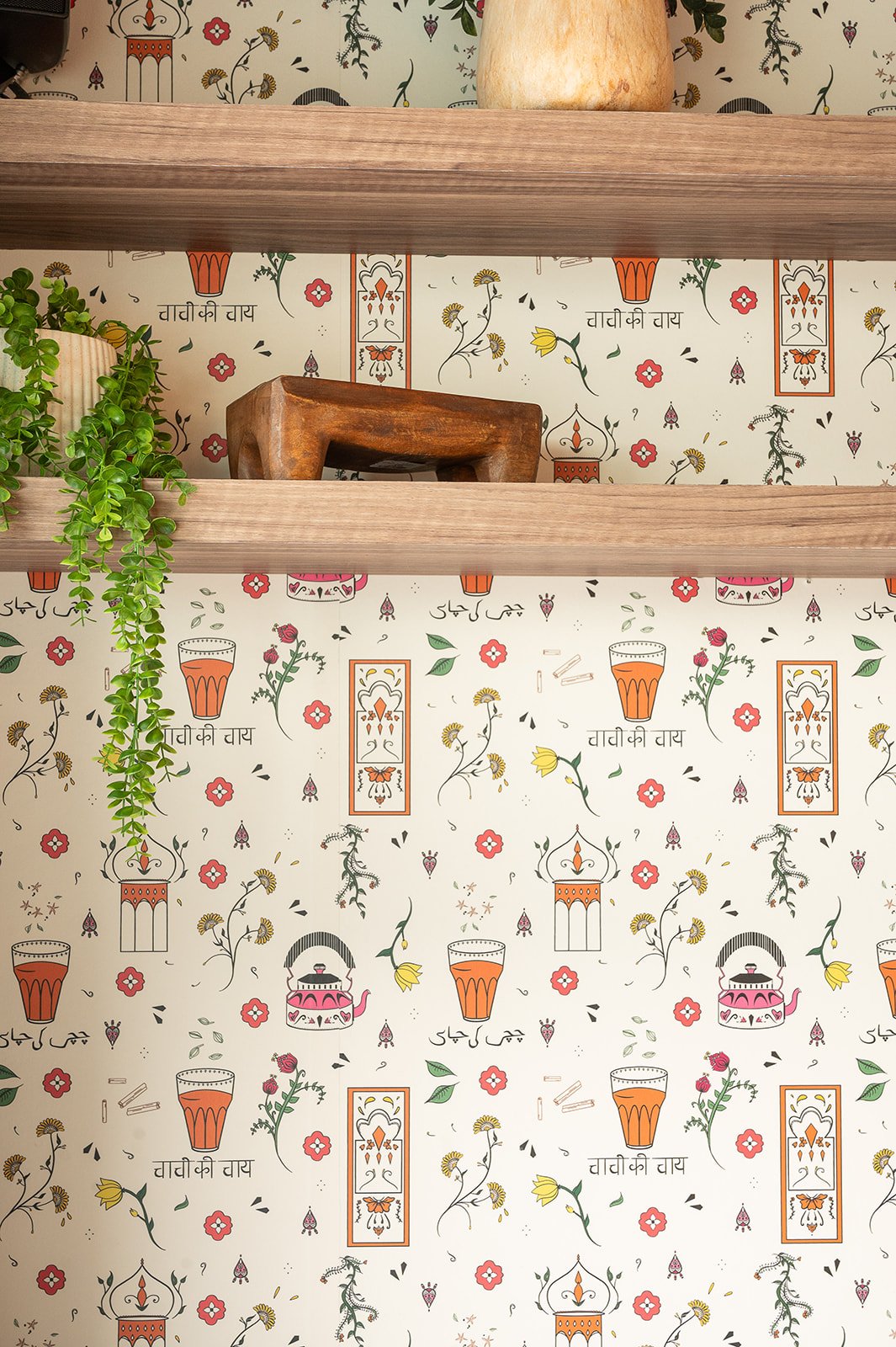 Close-up of Indian-inspired cafe wallpaper featuring pink teapots, orange chai glasses, floral designs, and Hindi script behind wooden floating shelves.