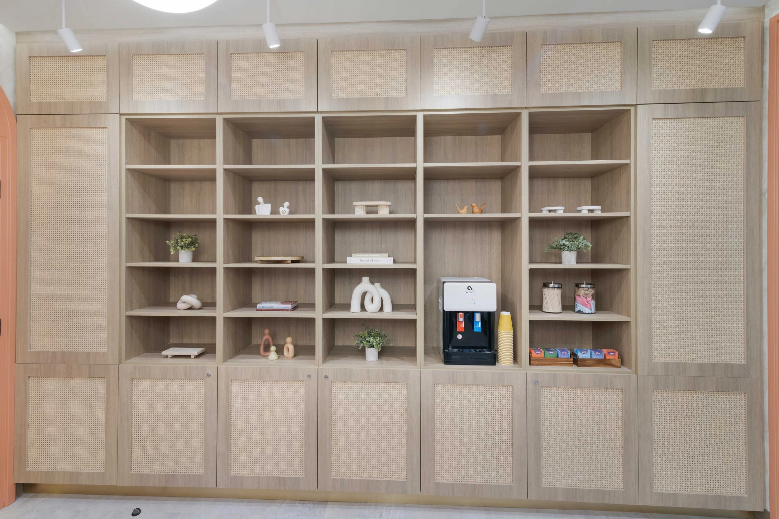 Custom light wood built-in shelving with rattan cabinet doors in the Aliso Dermatology waiting area, featuring decorative styling and a patient refreshment station.