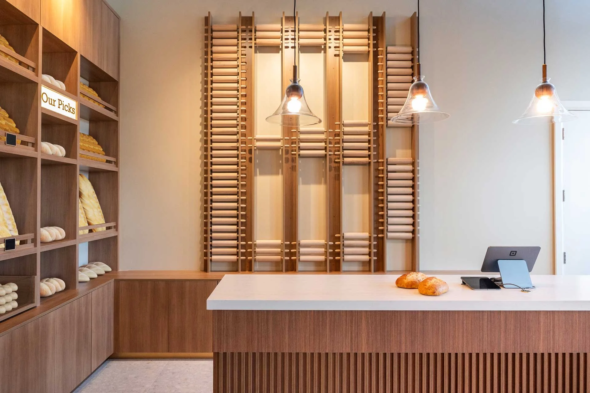 custom bakery check out counter with wood slats, quartz countertop, and glass pendants above, behind is a rolling pin design wall feature
