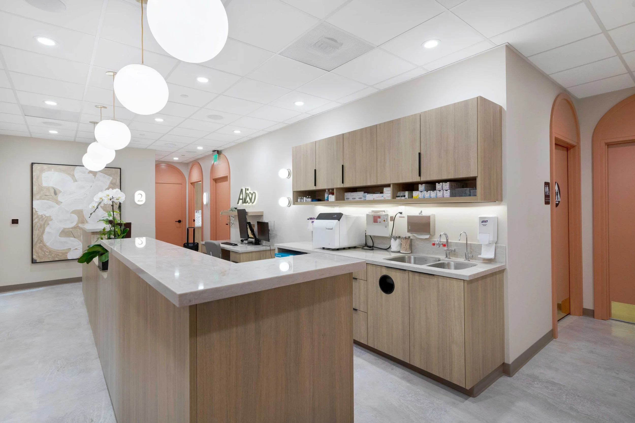 Interior hallway and staff station at Aliso Dermatology clinic showcasing a long wooden desk, globe pendant lighting, and signature peach arched exam room doors.