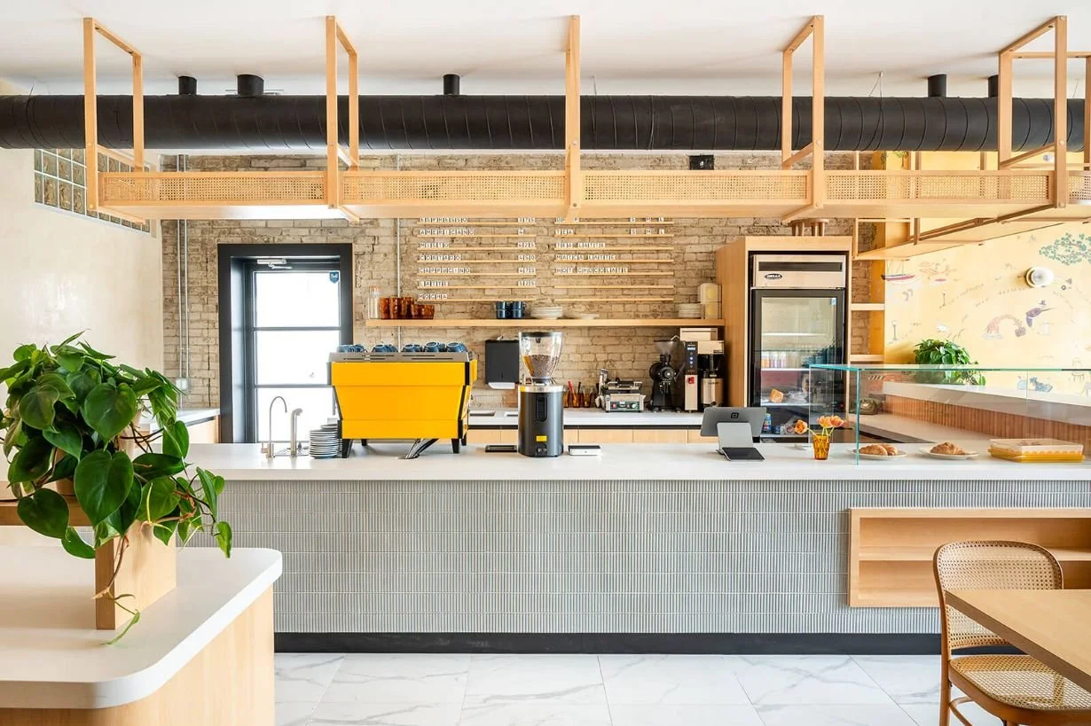 Cafe counter with blue small kit kat tiles completed with a white stone countertop, bright yellow espresso machine sits on top beside a pastry display case