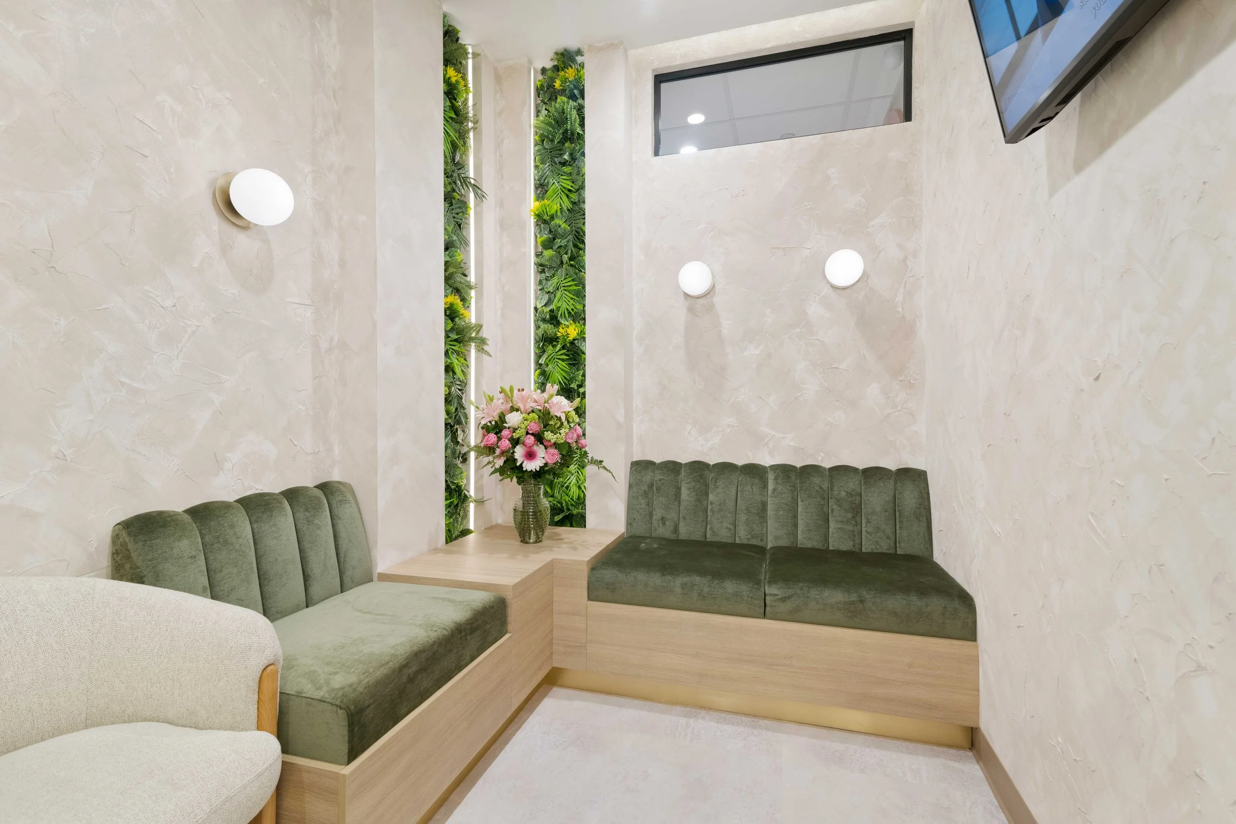 Cozy waiting room nook at Aliso Dermatology with green velvet bench seating, textured walls, a floral arrangement, and a vertical botanical wall accent.