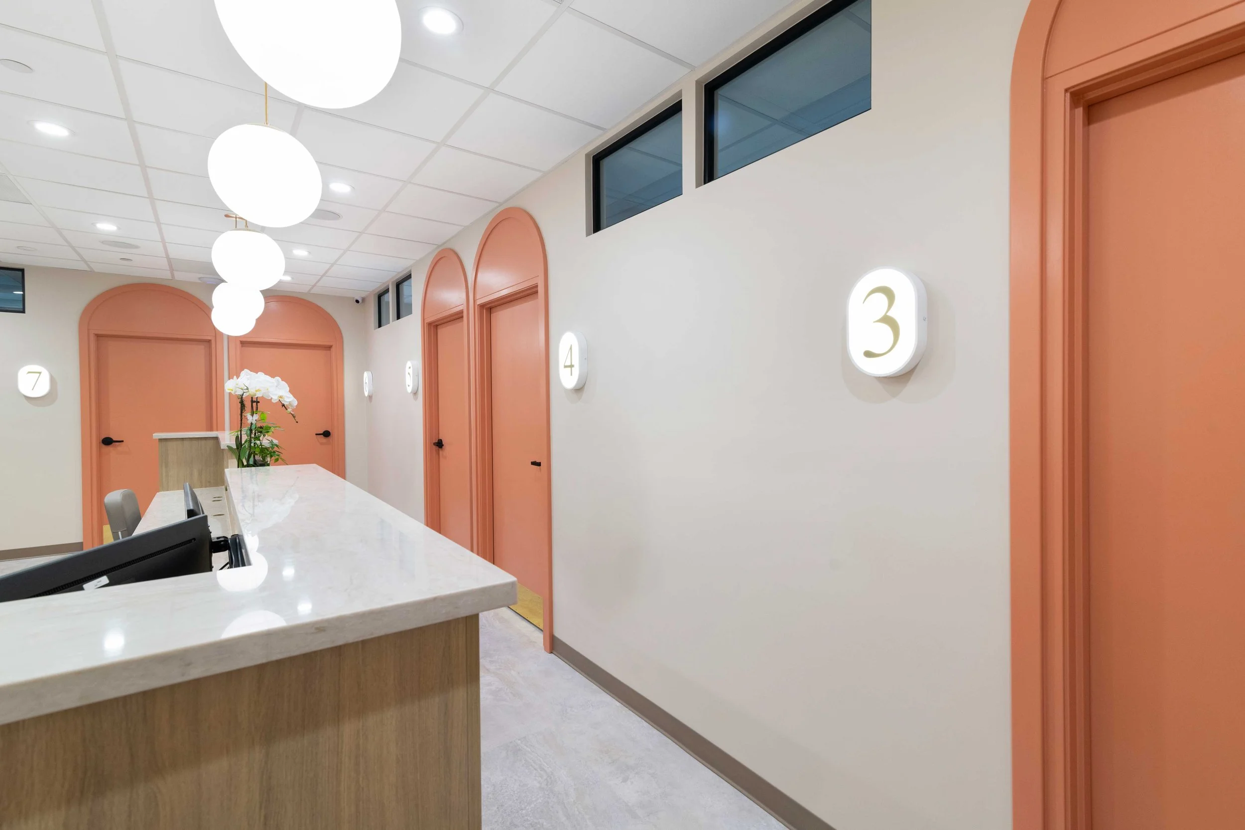 Bright dermatology clinic hallway featuring distinctive peach arched doors with illuminated room numbers and modern globe pendant lighting.