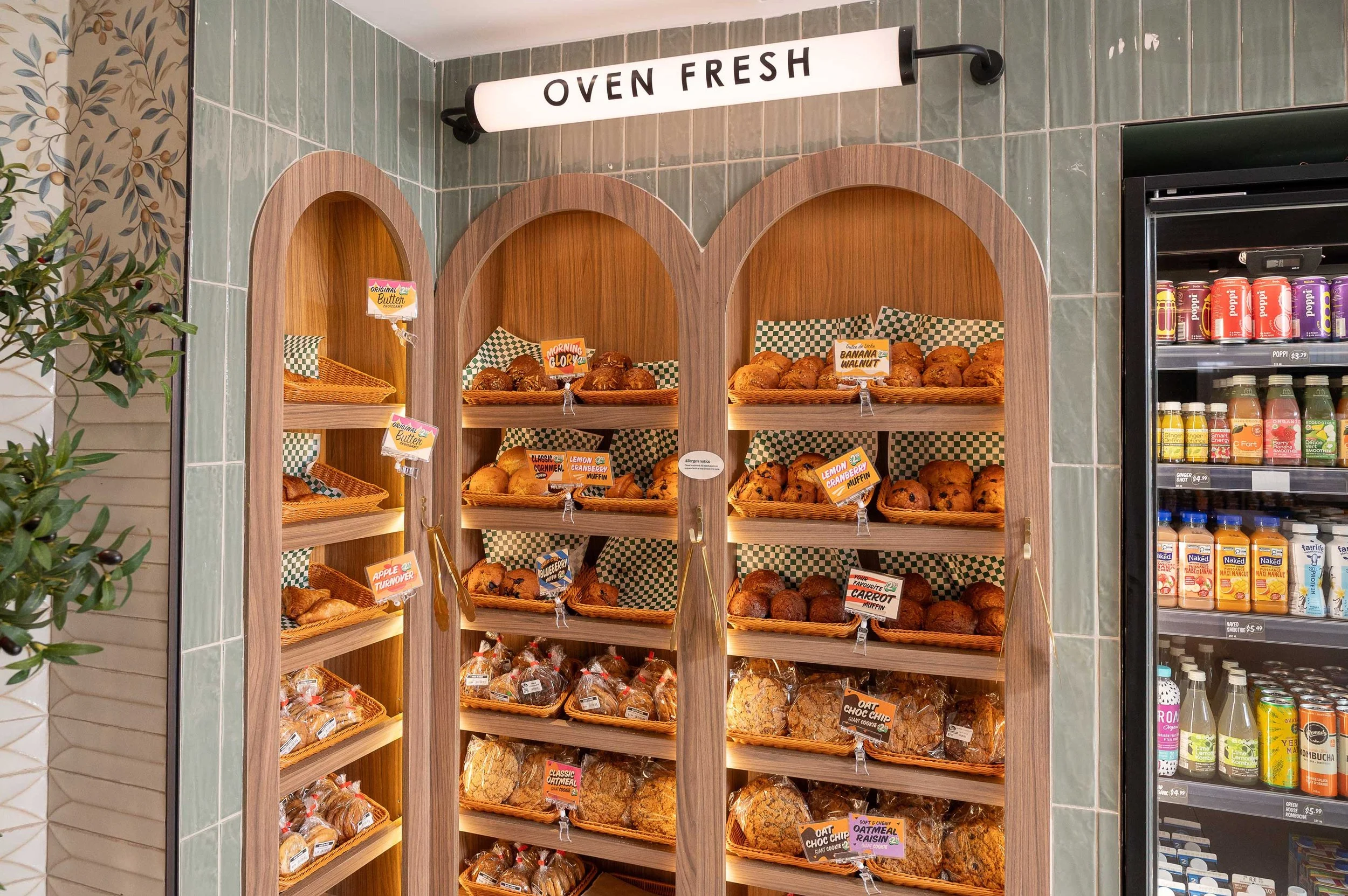 Light walnut arched bakery displays recessed into sage green tile wall with tubular illuminated signage above