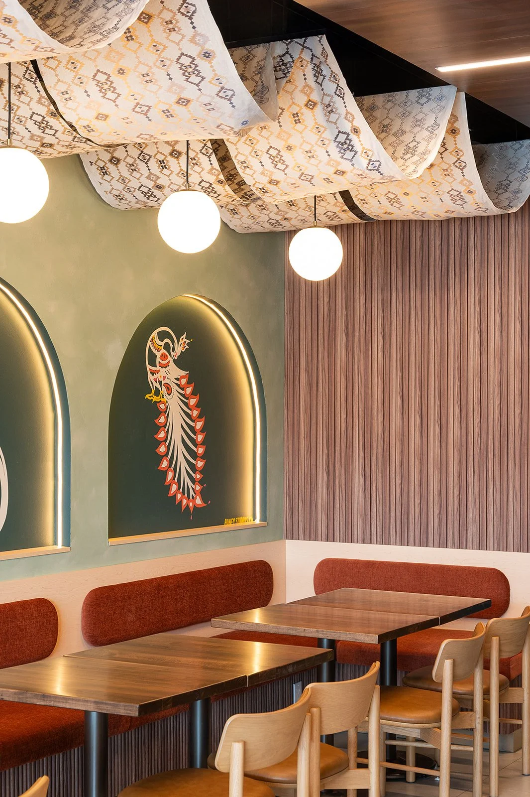 Corner booth in a modern cafe featuring a rust-orange banquette, wood slat walls, an illuminated peacock mural, and draped fabric ceiling decor.
