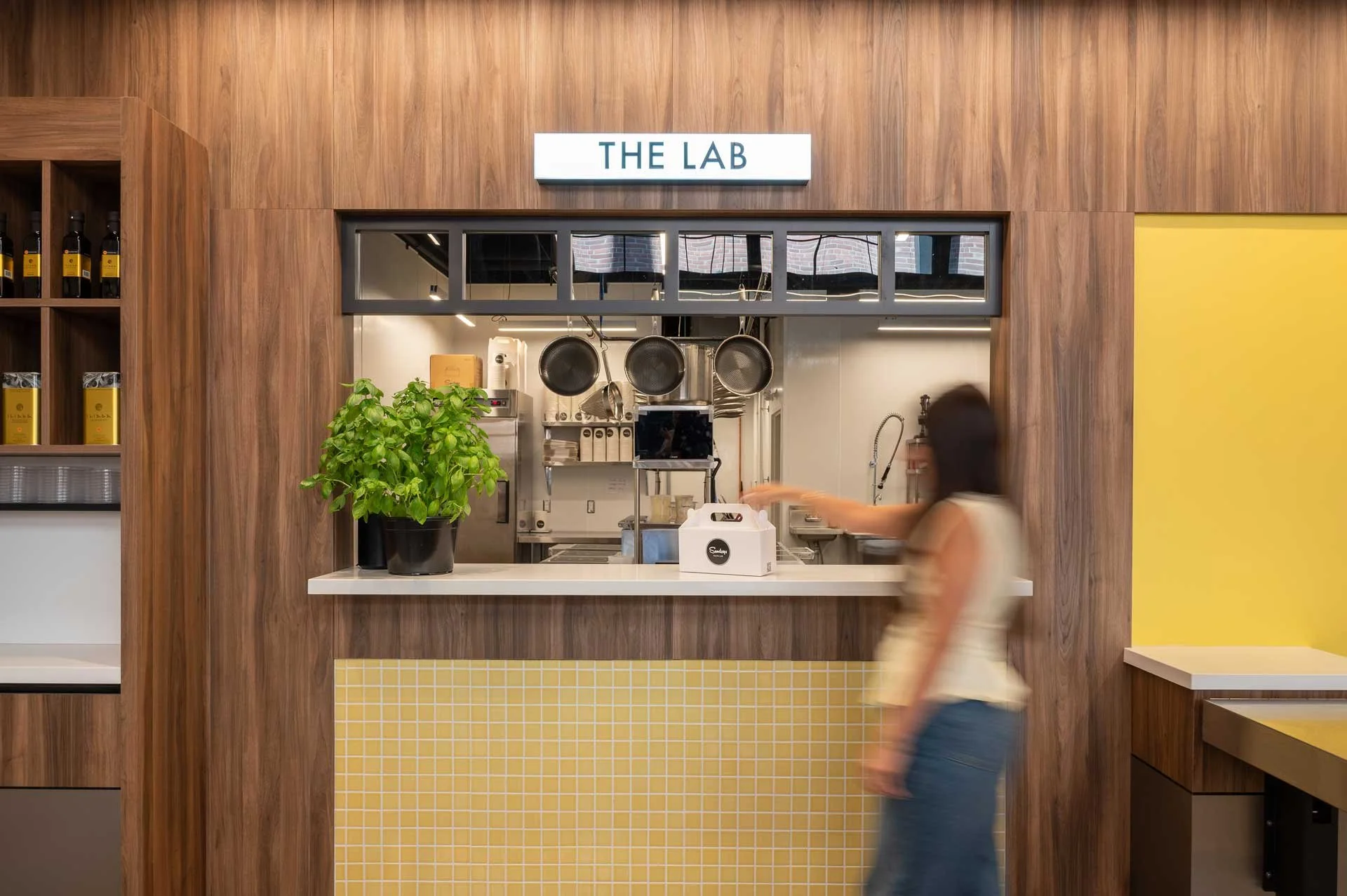 Restaurant passthrough window with illuminate THE LAB signage above. Wood walls and yellow mosaic tile with women grabbing food package