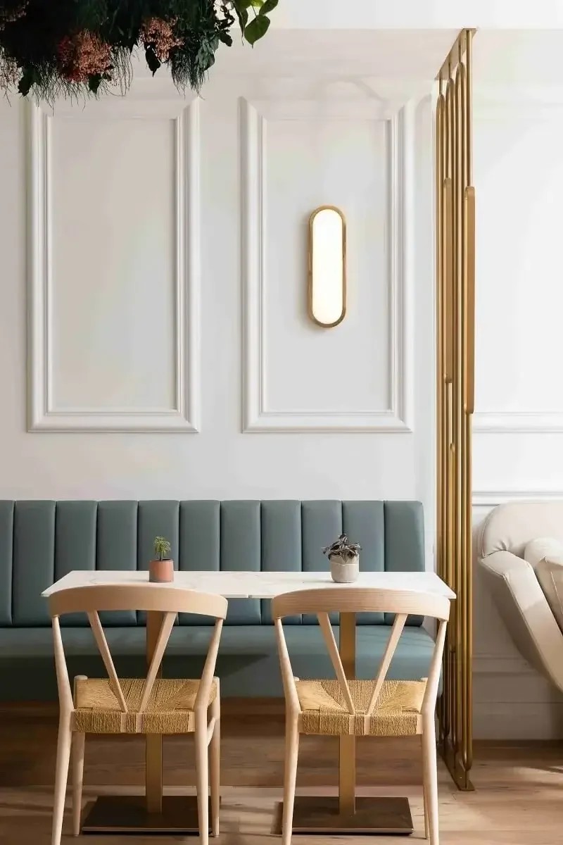 A stylish dining area with a white wall featuring decorative panels, a vertical wall light, a teal upholstered bench, two wooden chairs with woven seats, a white table with two small potted plants, and a decorative gold partition on the right side.