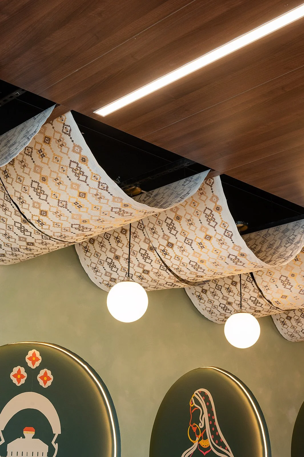 Cafe ceiling details showing patterned fabric drapes, modern brass globe pendant lights, and an arched mural of a hand holding a pink chai glass.
