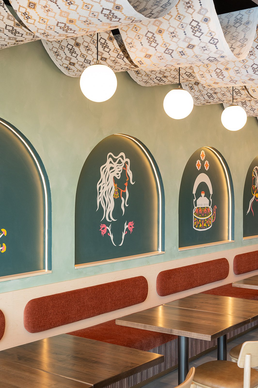 Indian-inspired cafe interior design showcasing a long orange banquette, wooden tables, hanging globe lights, and cultural wall murals inside archways.
