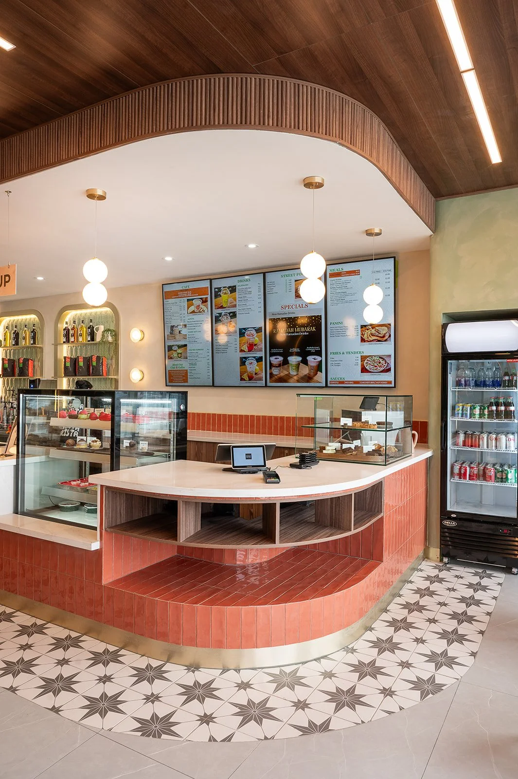 Modern cafe ordering counter featuring a curved orange tiled base, glass pastry display cases, digital menu boards on the wall, and decorative star-patterned floor tiles.