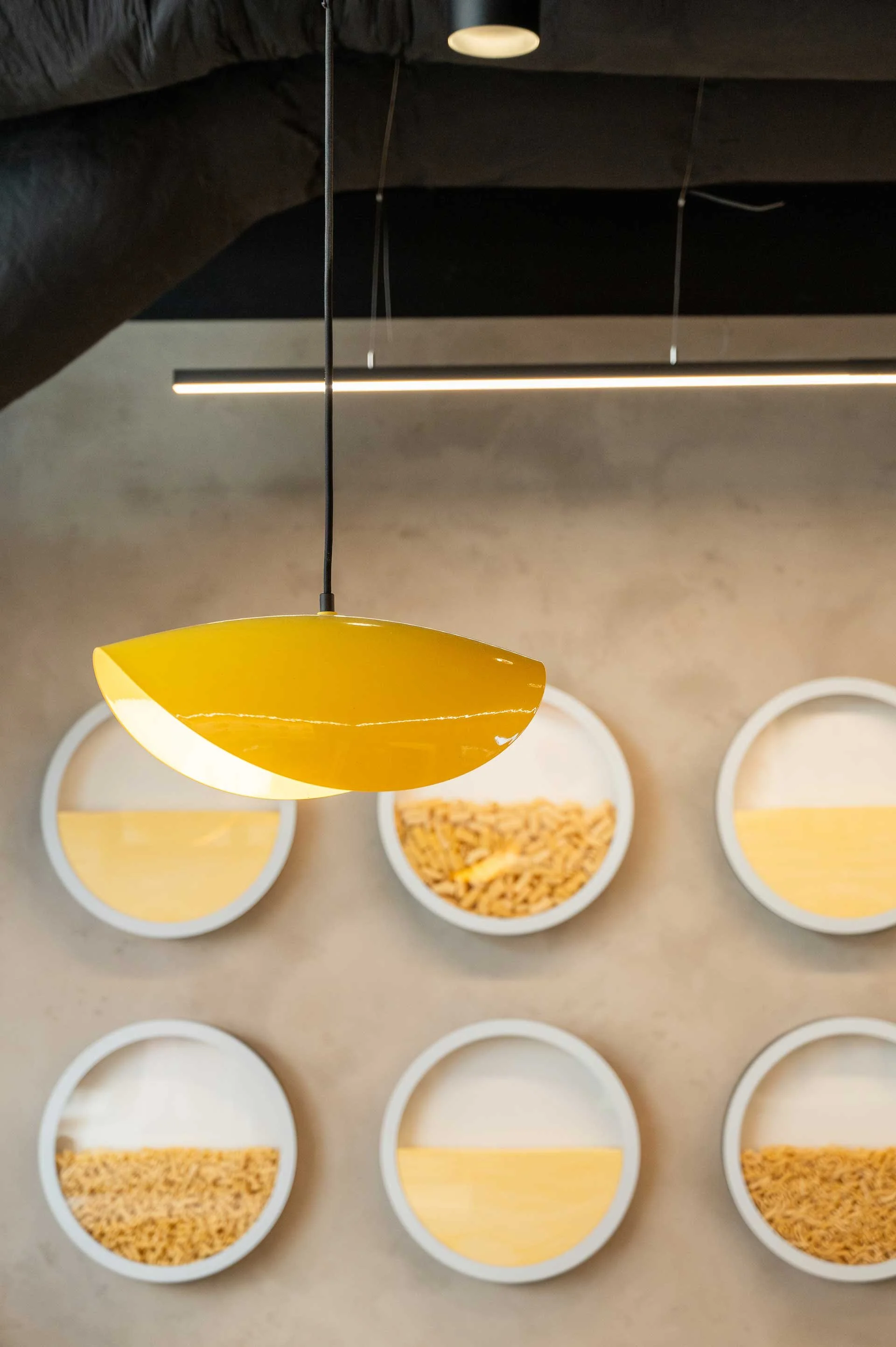 Yellow pasta noodle shaped pendant with a concrete wall behind and 6 circular pasta display