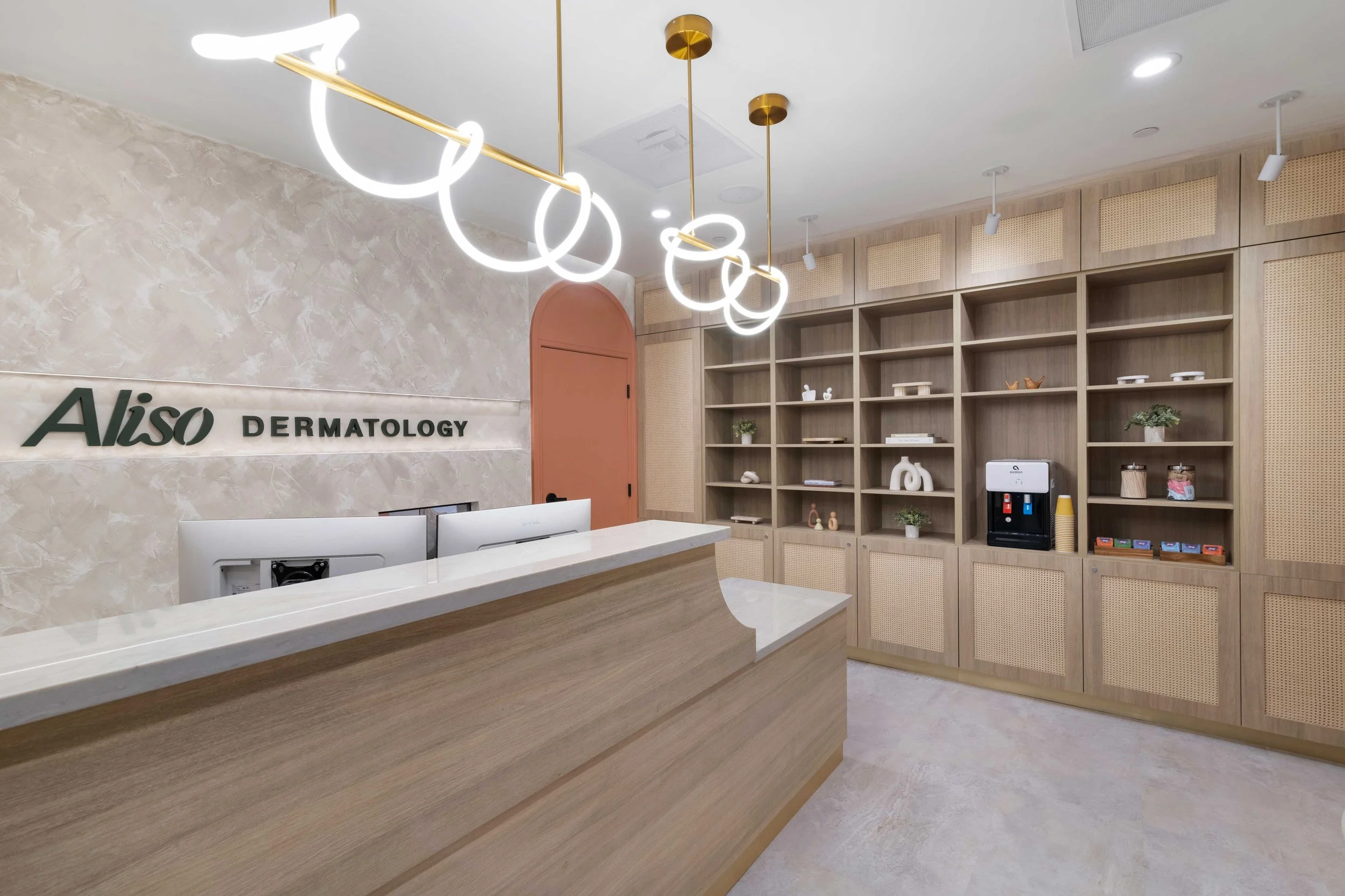 Aliso Dermatology reception desk featuring textured walls, custom wood and rattan shelving, and a modern continuous loop LED chandelier.