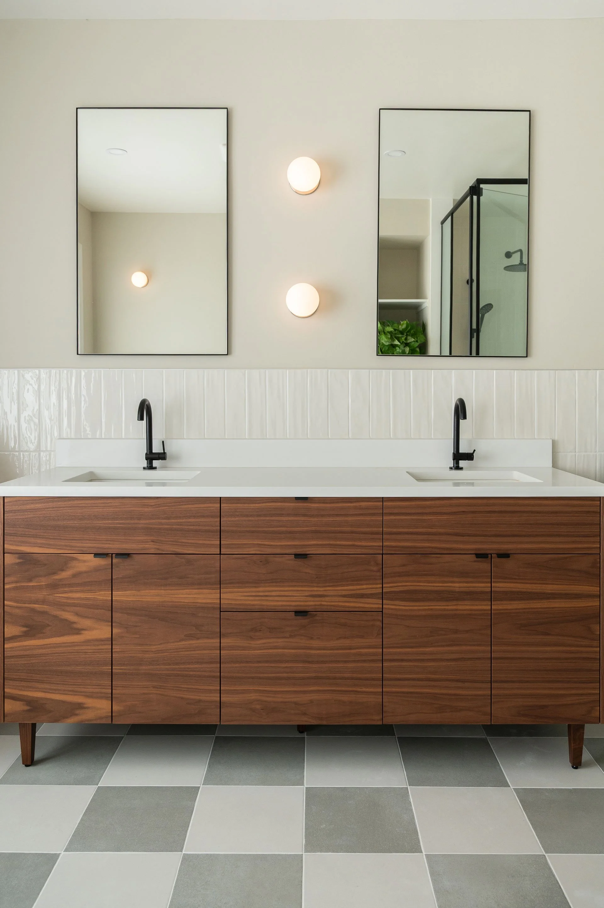 Doubled vanity with walnut wood cabinets on walnut furniture legs. White quartz countertop and matte black faucets. Large handmade tiles stacked vertically halfway up the wall with rectangular mirrors and Anony globe wall sconces