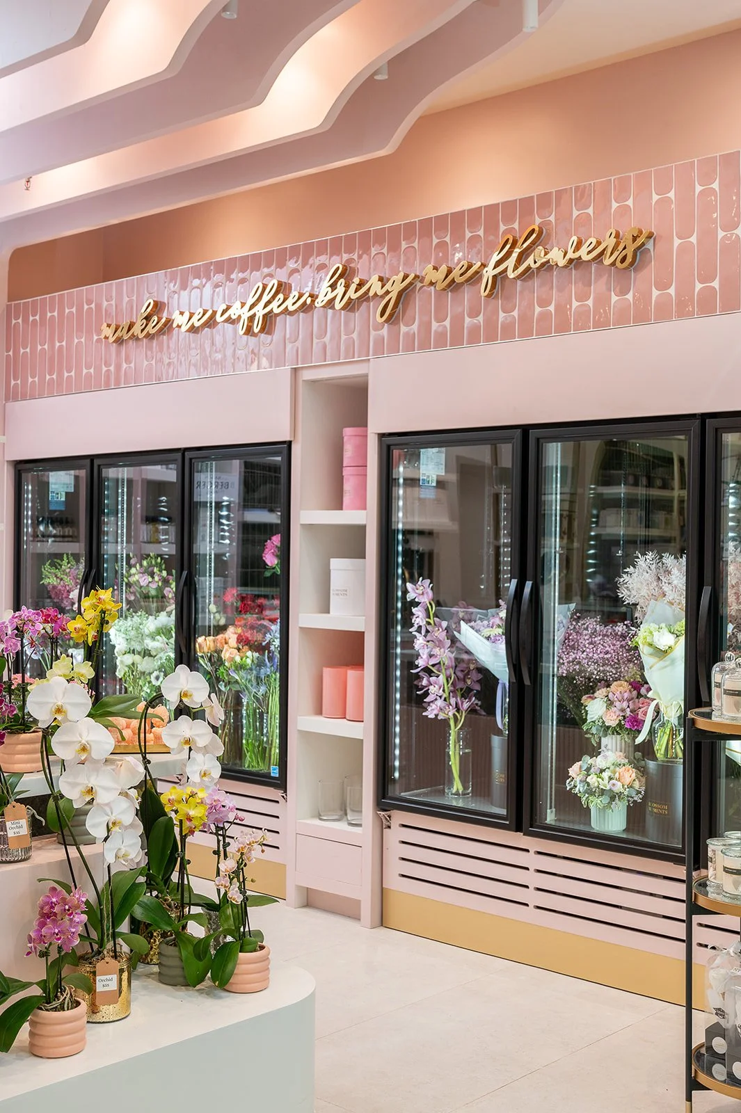 Gold script signage reading 'make me coffee, bring me flowers' on pink tile above the floral cooler display at Blossom Moments Scotia Plaza