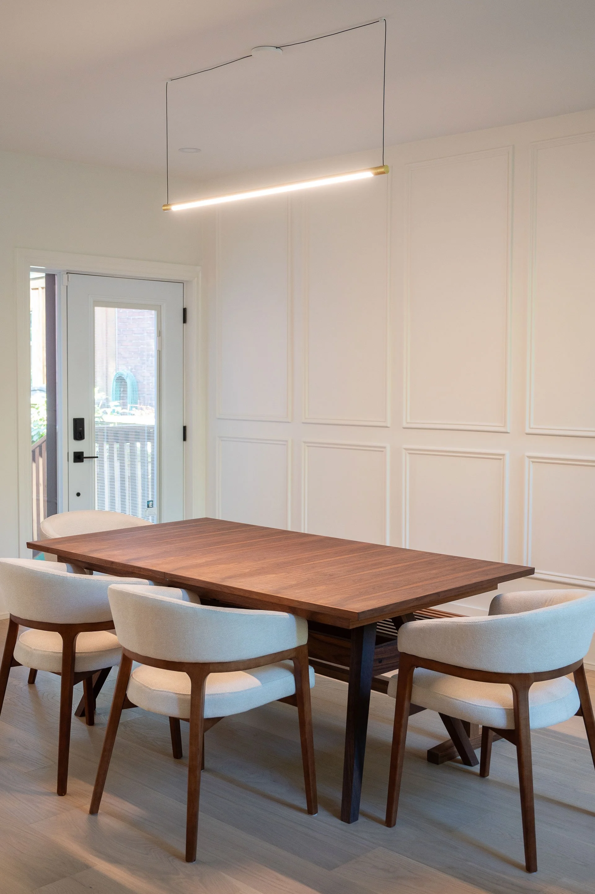 Walnut dining table with cream upholstered dining chairs around and linear anony  light fixture above. White wall paneling behind table