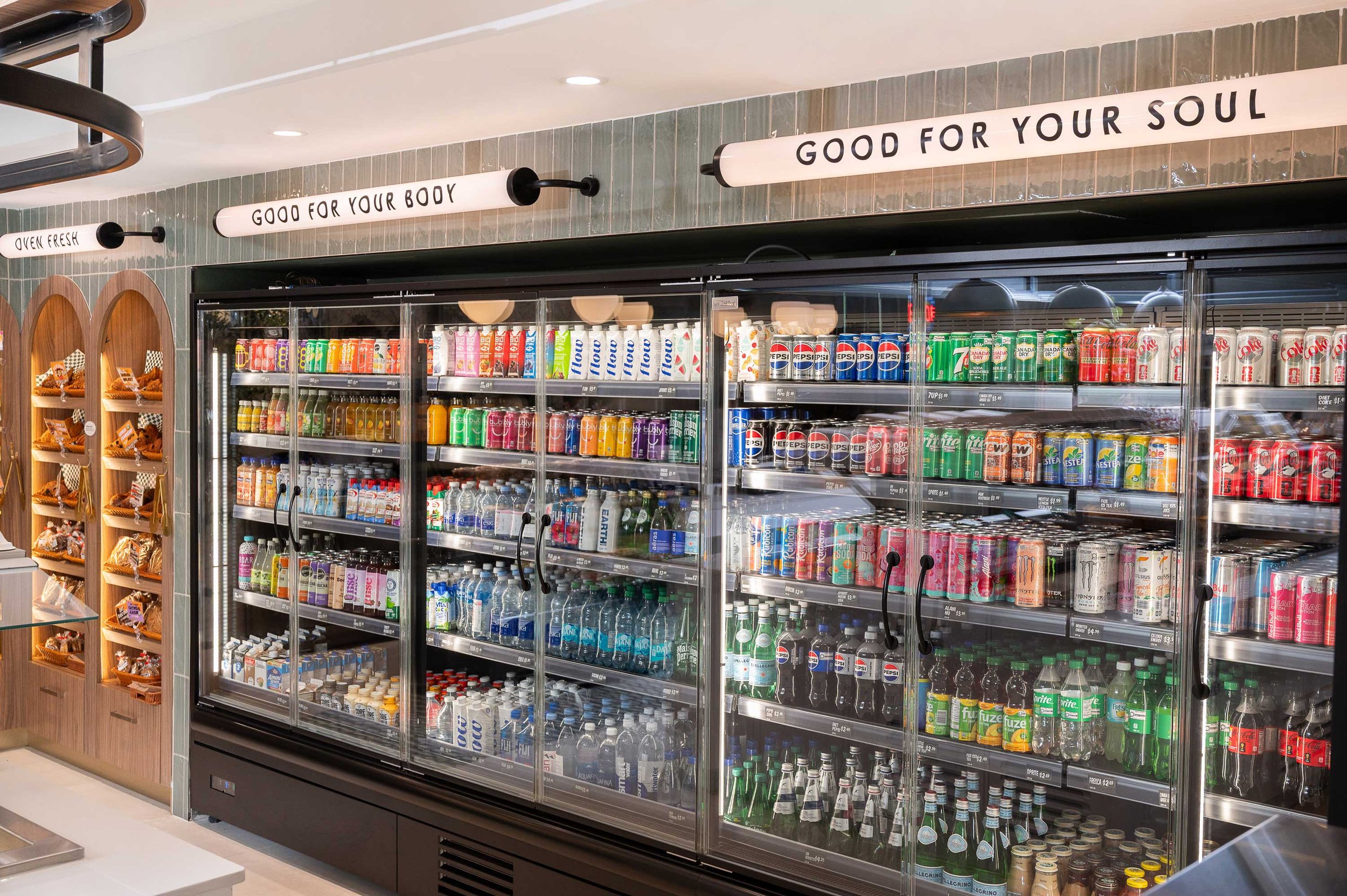 Display fridges recessed into sage green tiled wall with tubular illuminated signage above