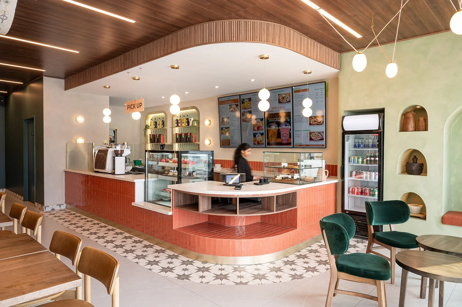 Wide view of a modern cafe interior featuring a curved orange tiled service counter, digital menu boards, pastry display cases, and dining areas with green velvet chairs and wooden tables.