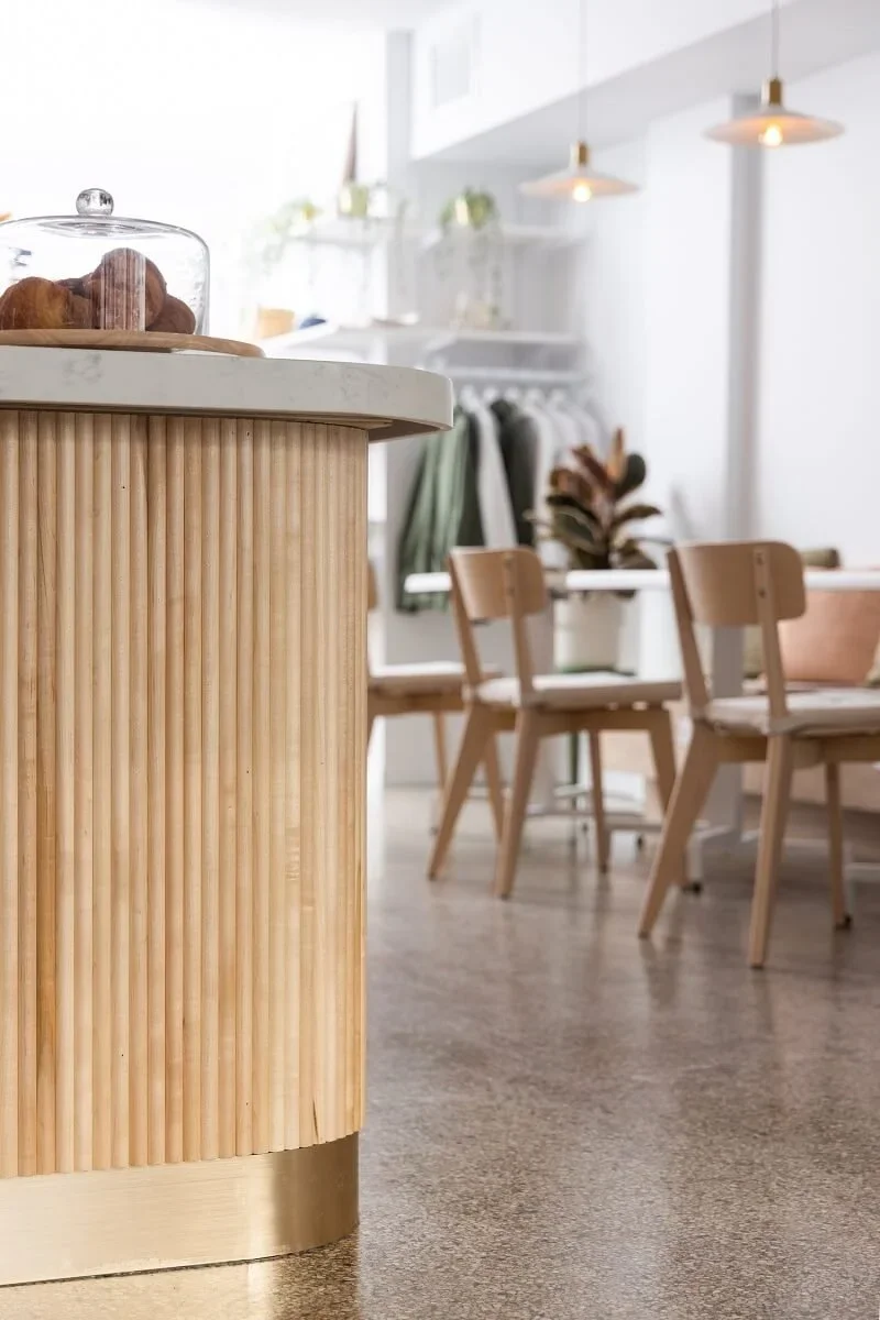 A modern kitchen with a wooden rounded island counter, a glass dome covering baked goods, a dining table with wooden chairs, and hanging pendant lights.
