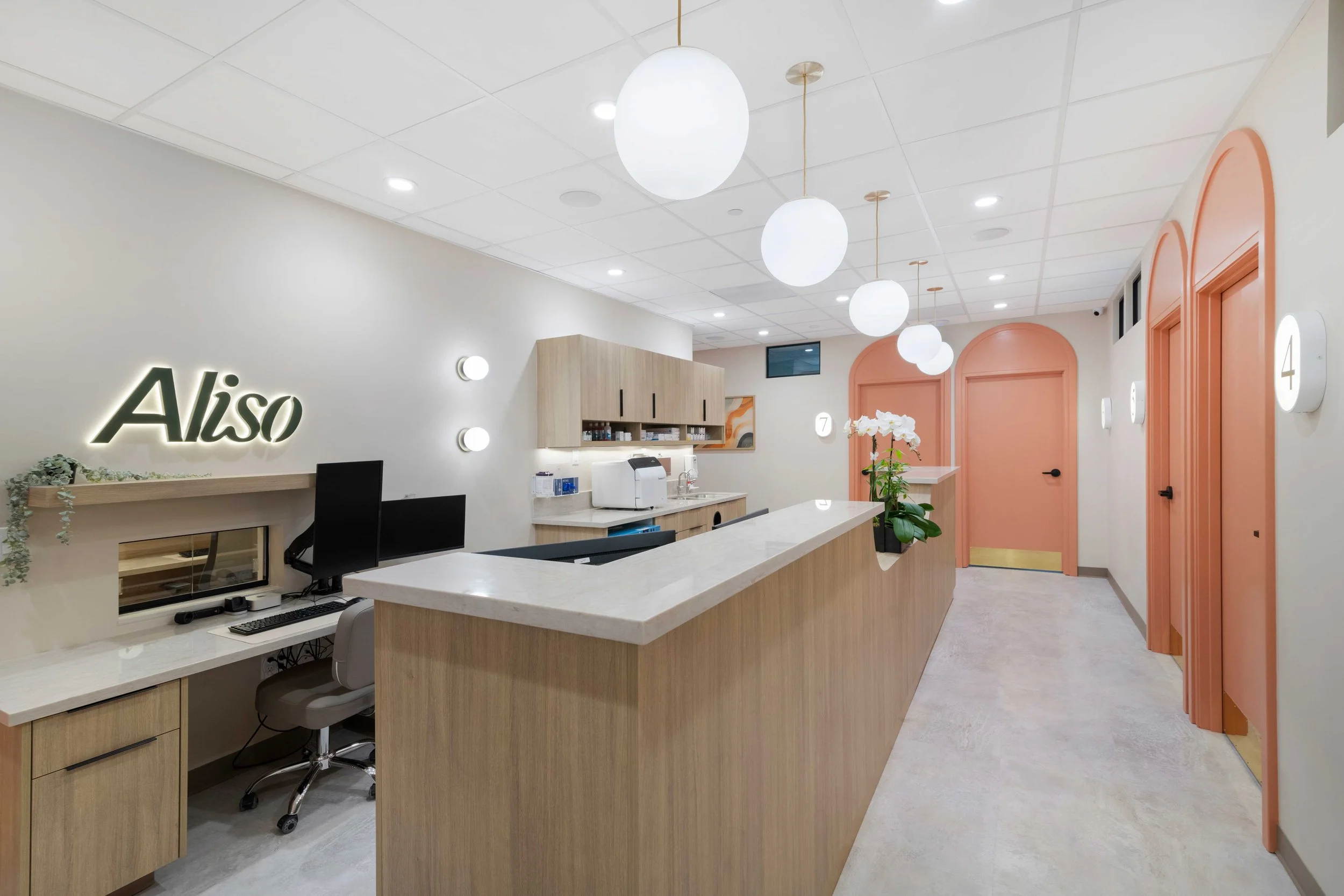 Interior hallway and staff station at Aliso Dermatology clinic showcasing a long wooden desk, globe pendant lighting, and signature peach arched exam room doors.