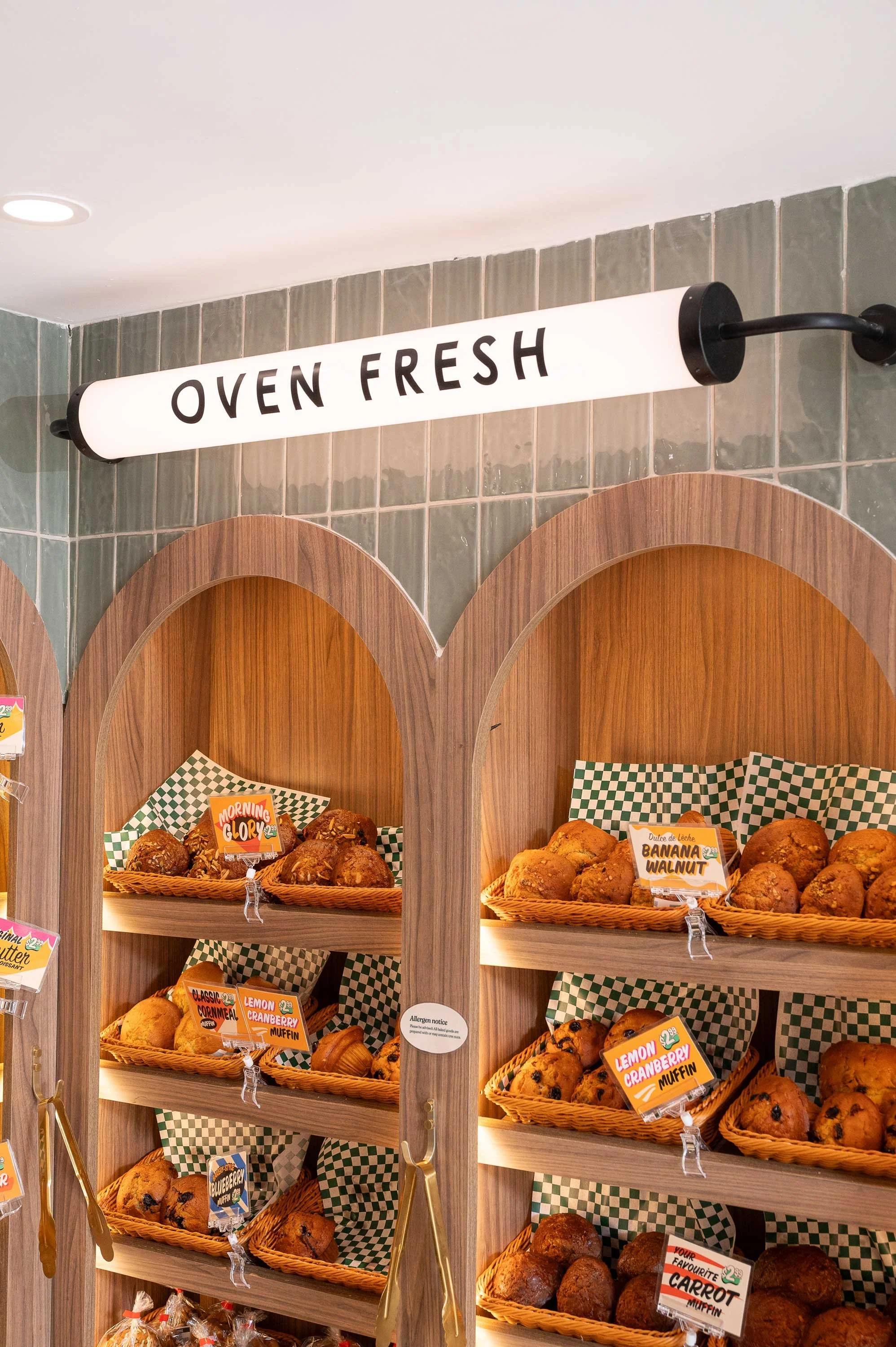 Light walnut arched bakery displays recessed into sage green tile wall with tubular illuminated signage above