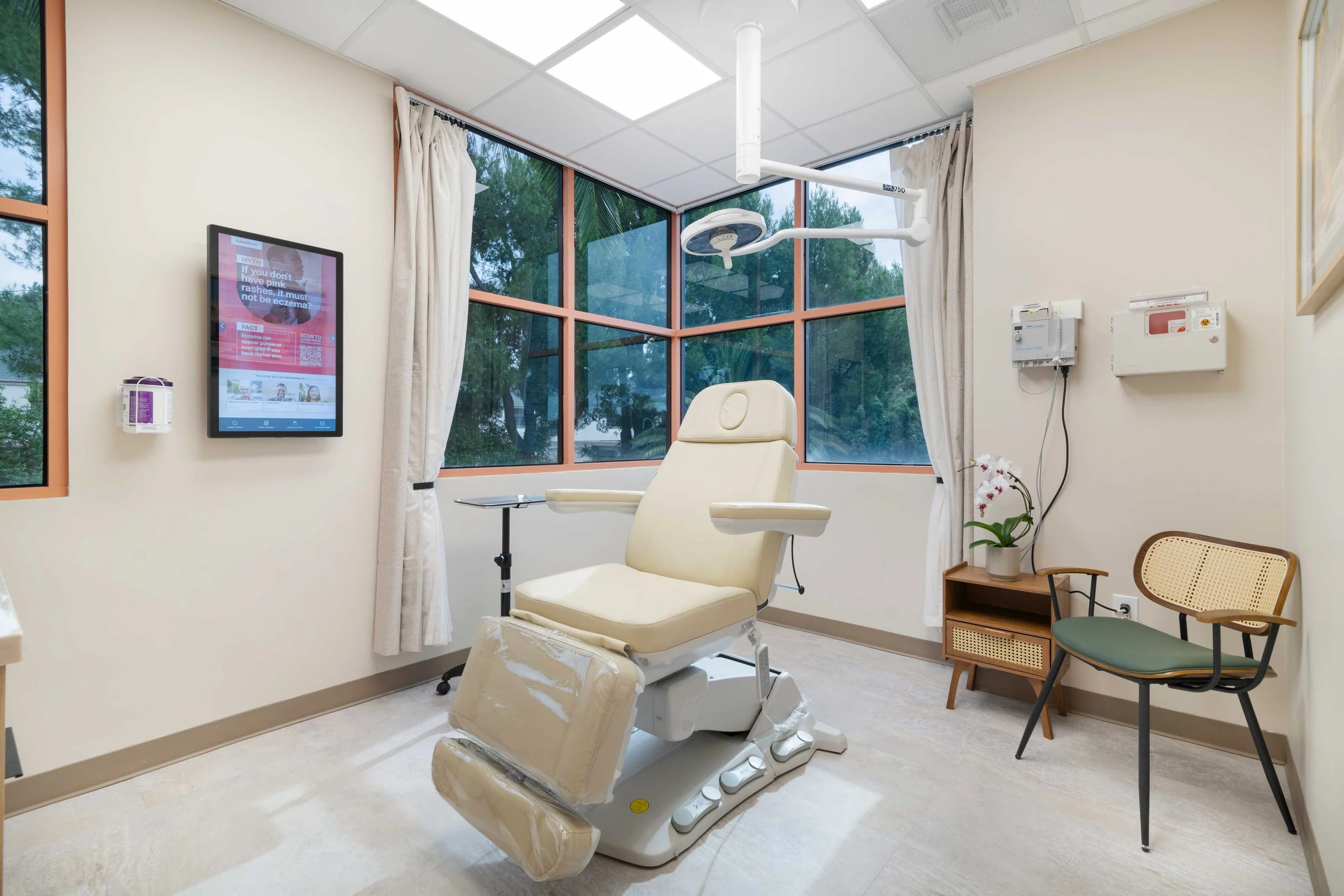 Bright, modern dermatology exam room at Aliso Dermatology with a beige patient treatment chair, surgical lighting, and large windows