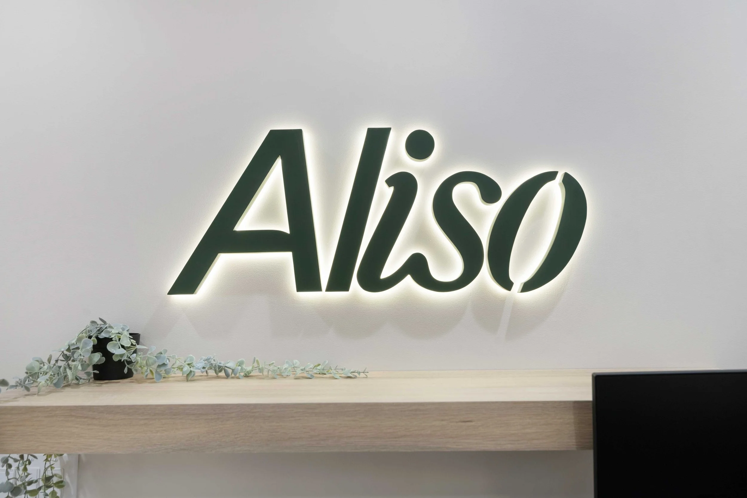 Backlit Aliso Dermatology logo sign on a clean white wall above a modern wooden counter with decorative green trailing plants.