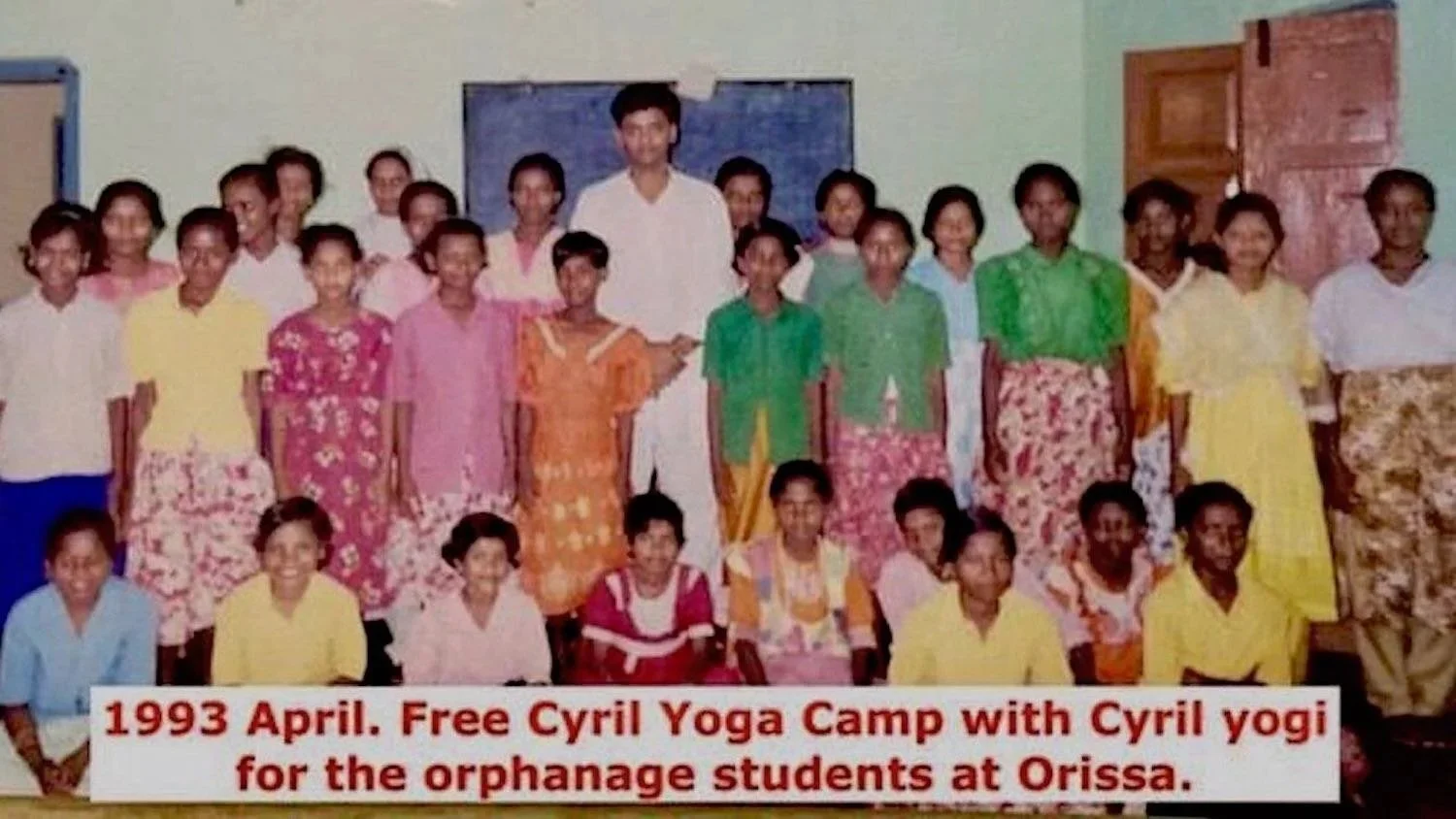 A group of children and a young man, likely Cyril Yogi, posing together in a classroom setting. The children are dressed in colorful clothing, and the background shows a blackboard and classroom door. The photo is from April 1993, with a caption indi
