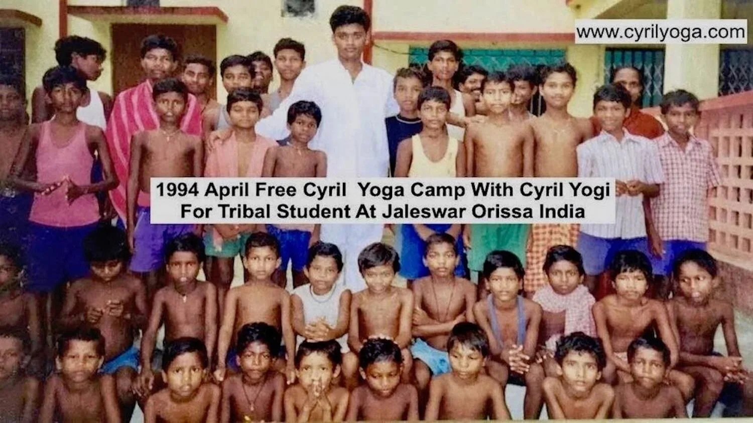 Group of children and a man in front of a yellow building, some children wearing traditional Indian clothing, participating in a 1994 Cyril Yoga camp in Jaleswar, Orissa, India.