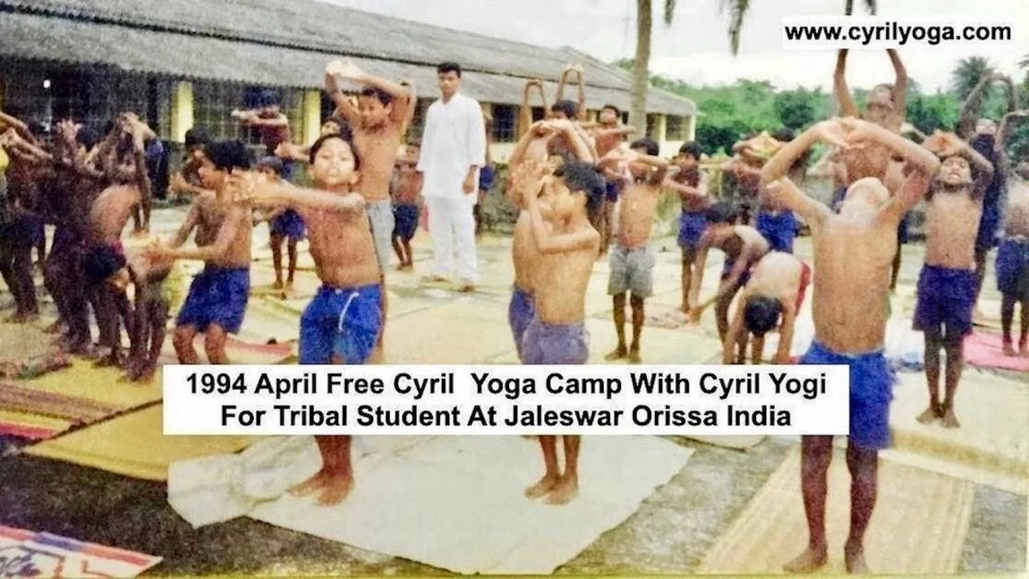 A large group of children and a few adults participating in a yoga event outdoors, with some children in a yoga pose and others waiting or looking around. The setting appears to be a school or community area with a building in the background. The ima