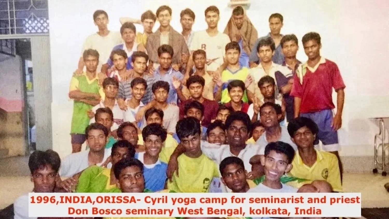 A group of young boys and a few adults gathered together indoors, posing for a group photo at Cyril yoga camp in 1996, near Kolkata, India.