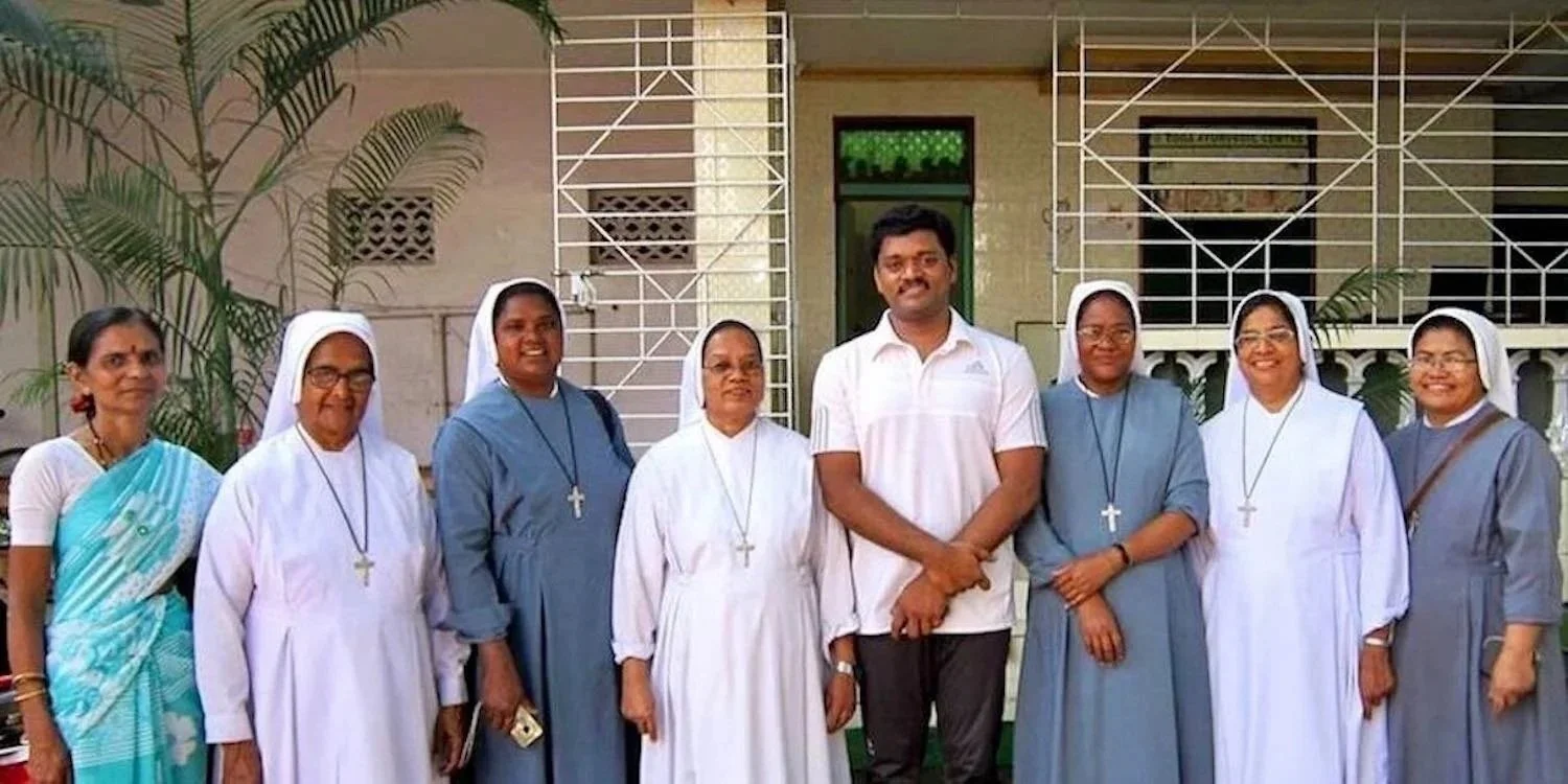 Group of eight women in religious habits and a man in a white polo shirt standing outdoors in front of a building with plants.