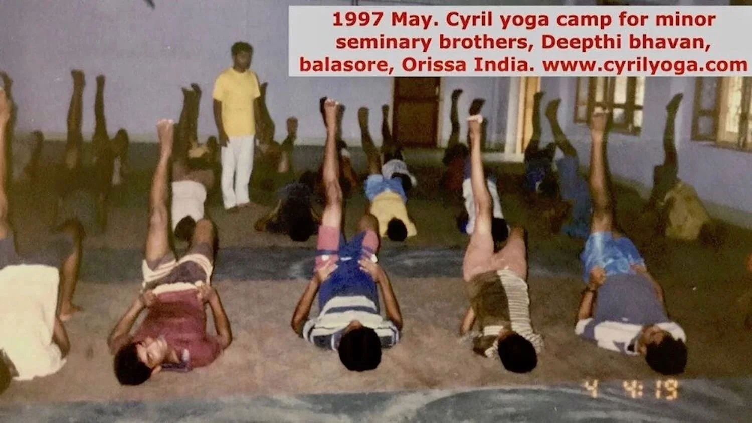 Children practicing yoga in a classroom during a camp in India in 1997, with a teacher overseeing them.