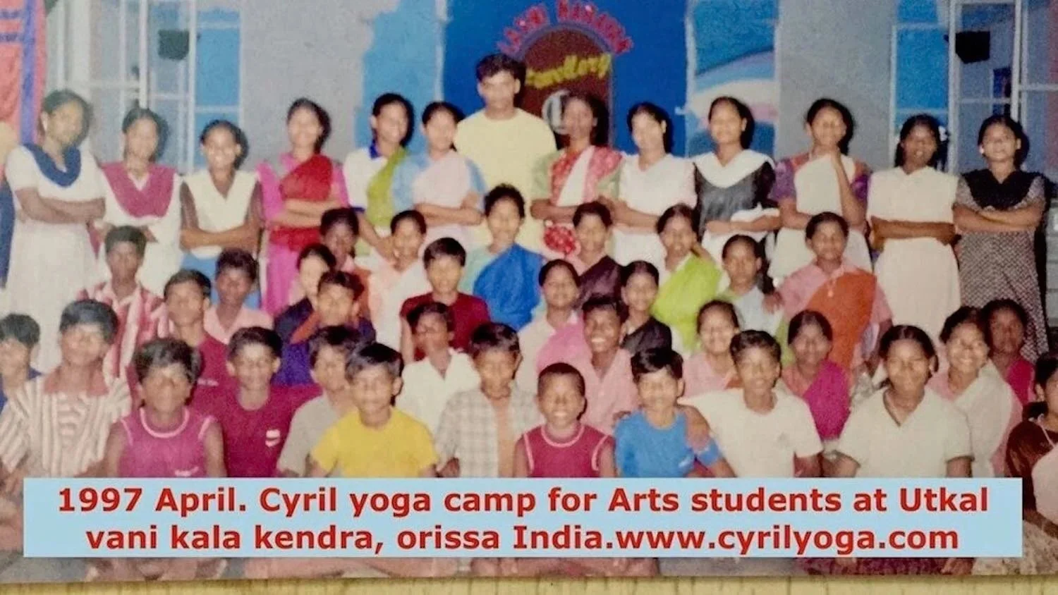 A large group of children and adults posing together on a stage in front of colorful decorations, attending a 1997 Cyril yoga camp for arts students at Utkal Vani Kala Kendra in Odisha, India.