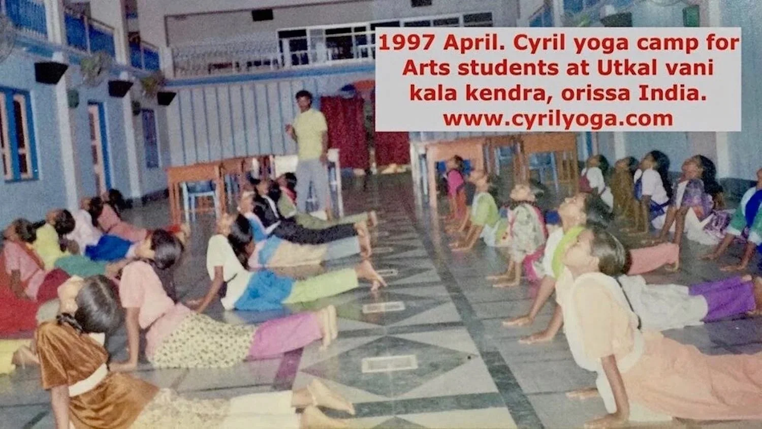 Children participating in a yoga camp, performing downward dog pose on the floor inside a room, with a male instructor standing nearby. A sign mentions a 1997 April Cyril yoga camp in Utkal Vani Kendra, Orissa, India.