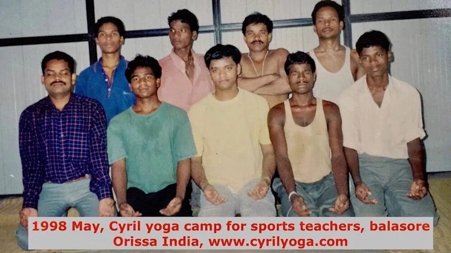 Group of nine men posing together, most making a meditative hand gesture, in a room with metal wall panels, during a yoga camp in May 1998 at Cyril yoga camp, Balasore, Orissa, India.
