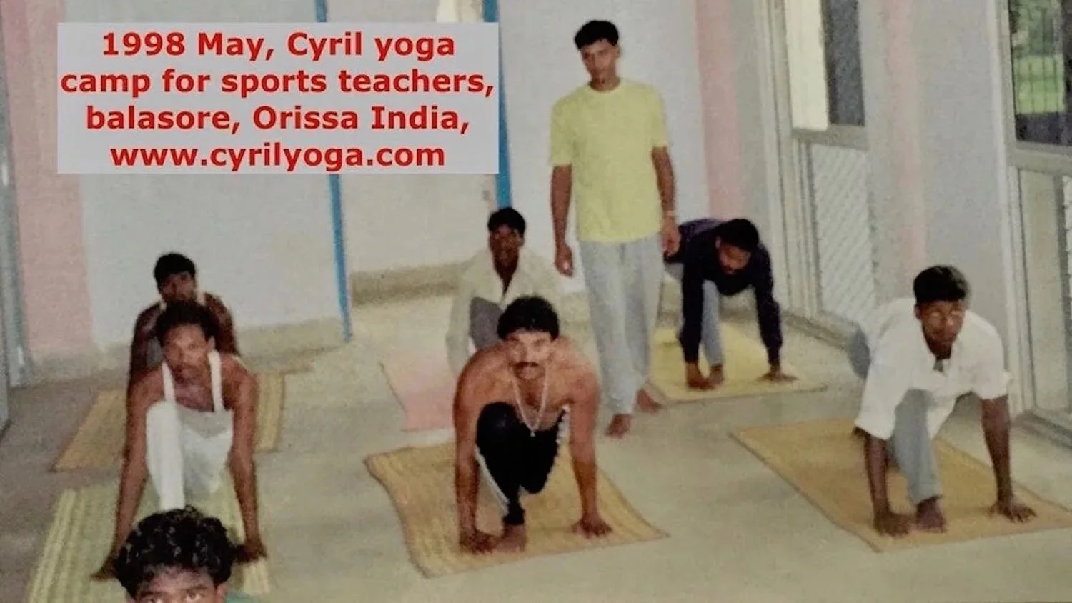 A group of six people, including a shirtless man, is practicing yoga on mats inside a room with large windows. One person is standing, while the others are in a downward-facing dog pose. There is a red text overlay at the top left corner of the image