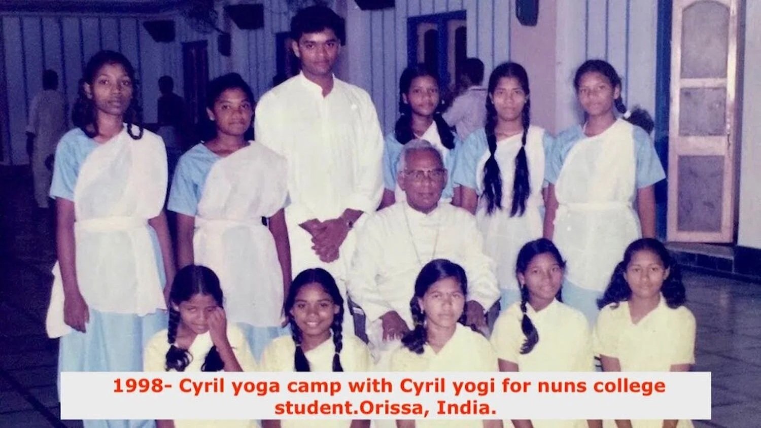 A group of twelve young girls and two men, one elderly and one younger, posing indoors in a room with wooden floors and walls. The girls are wearing light blue and white uniforms with braids, and the men are dressed in white. The photo has a caption 