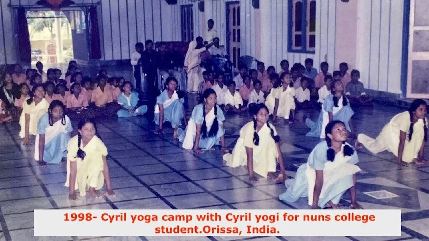 A group of young girls in light-colored dresses practicing yoga in a large indoor hall with wooden floors, supervised by an instructor. The text overlay indicates this is from 1998, at a Cyril yoga camp for nuns' college students in Orissa, India.