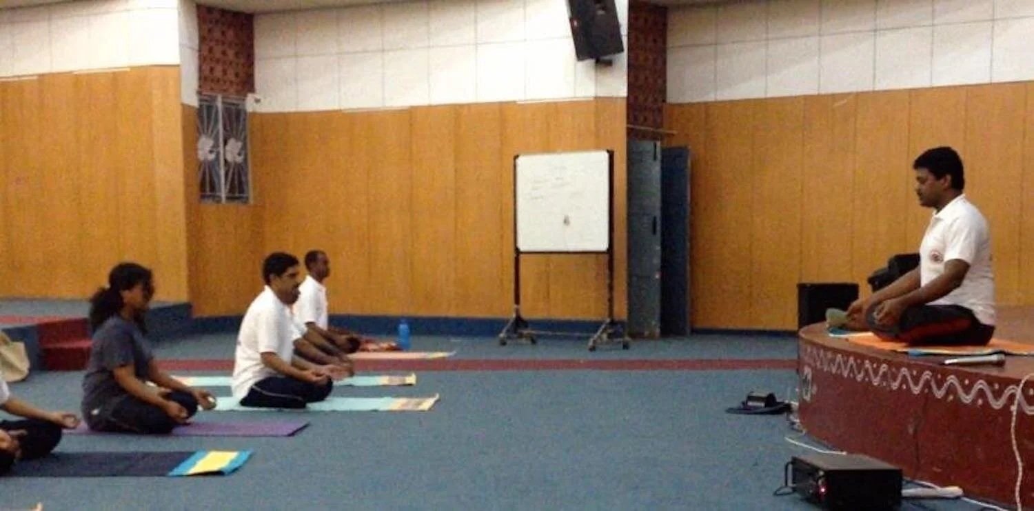 A group of individuals practicing meditation or yoga, seated cross-legged on mats, facing a person at a front platform in a room with wood-paneled walls and a whiteboard.